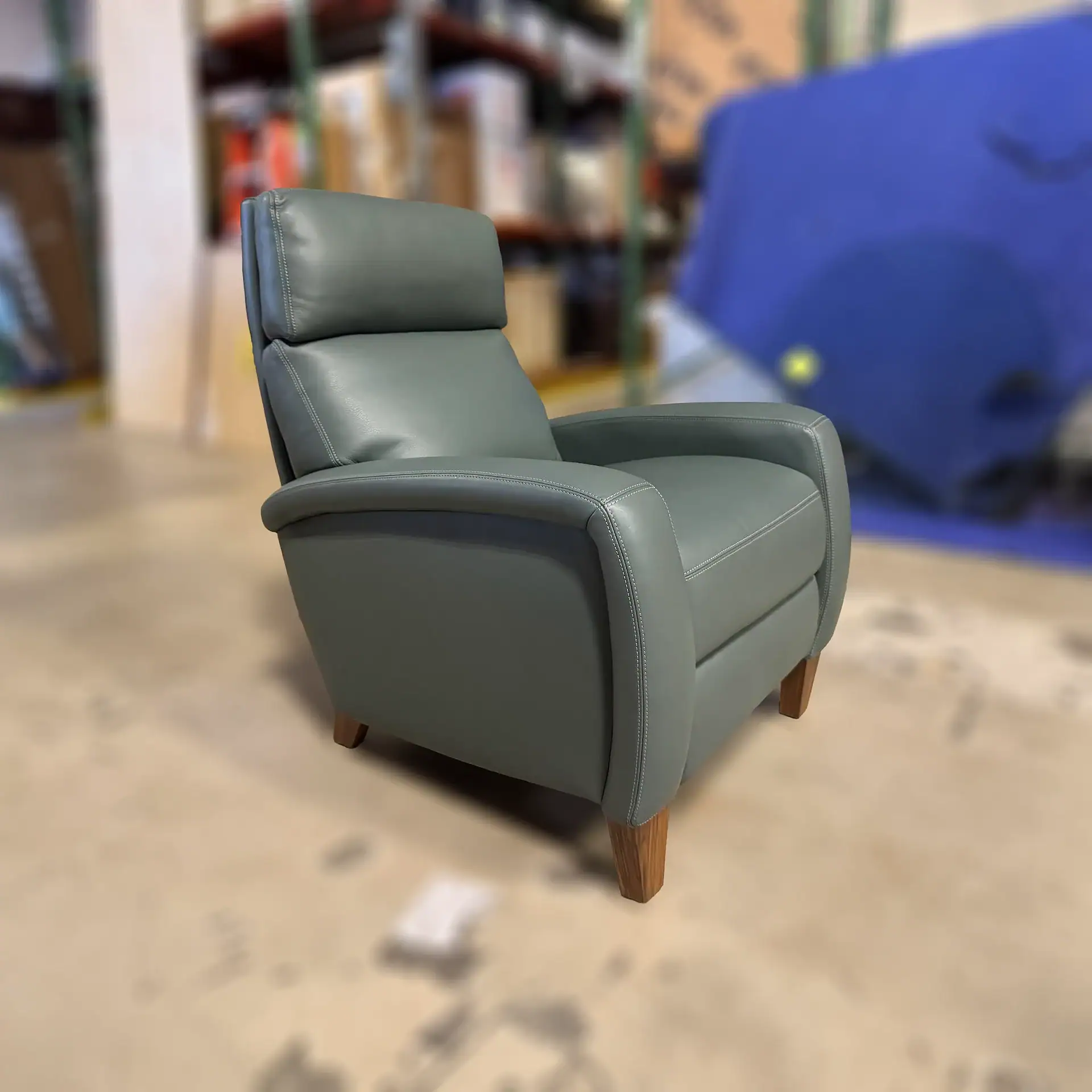 A sage green leather recliner chair with white contrast stitching and wooden legs, set against a blurred warehouse background.