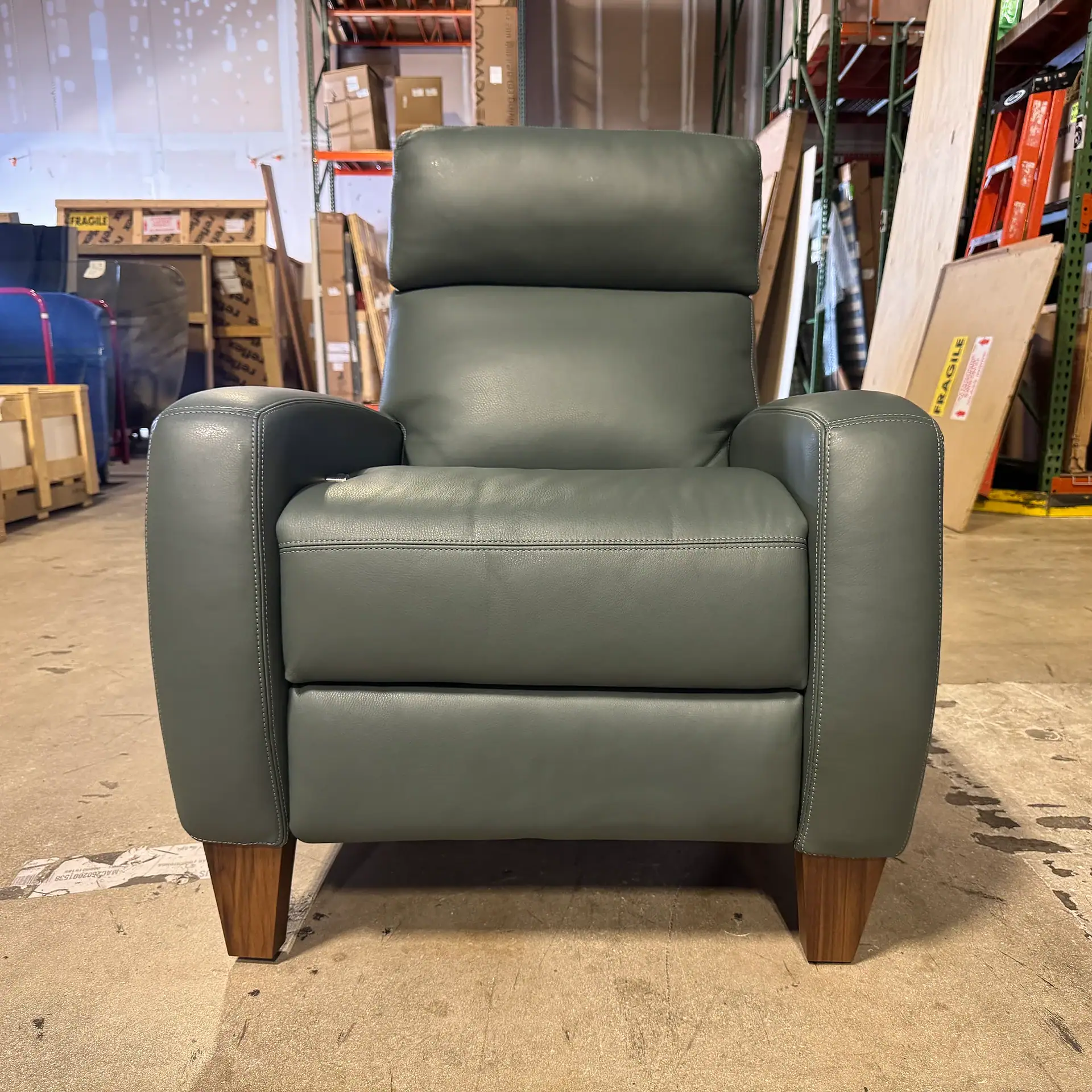 A dark gray leather recliner chair with wooden legs, centered in a warehouse setting with storage racks and boxes in the background.