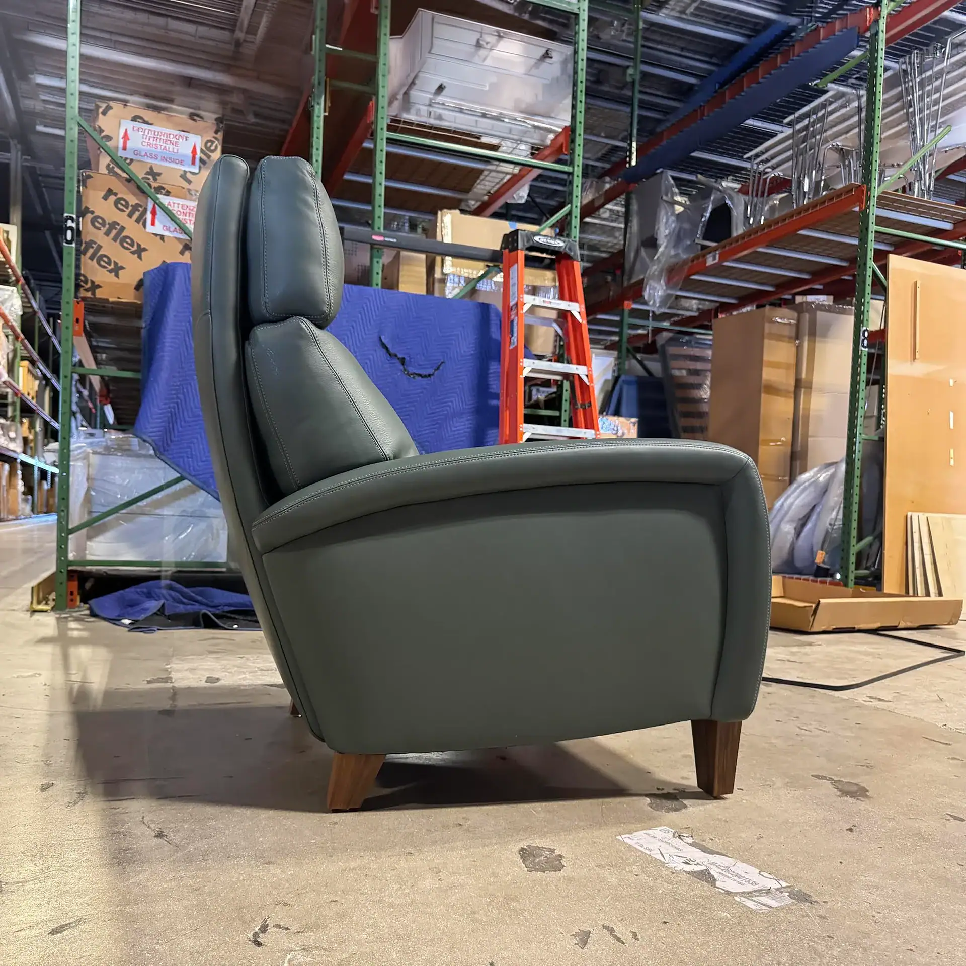 A dark green leather armchair with wooden legs in a warehouse, surrounded by shelving, boxes, and an orange ladder.