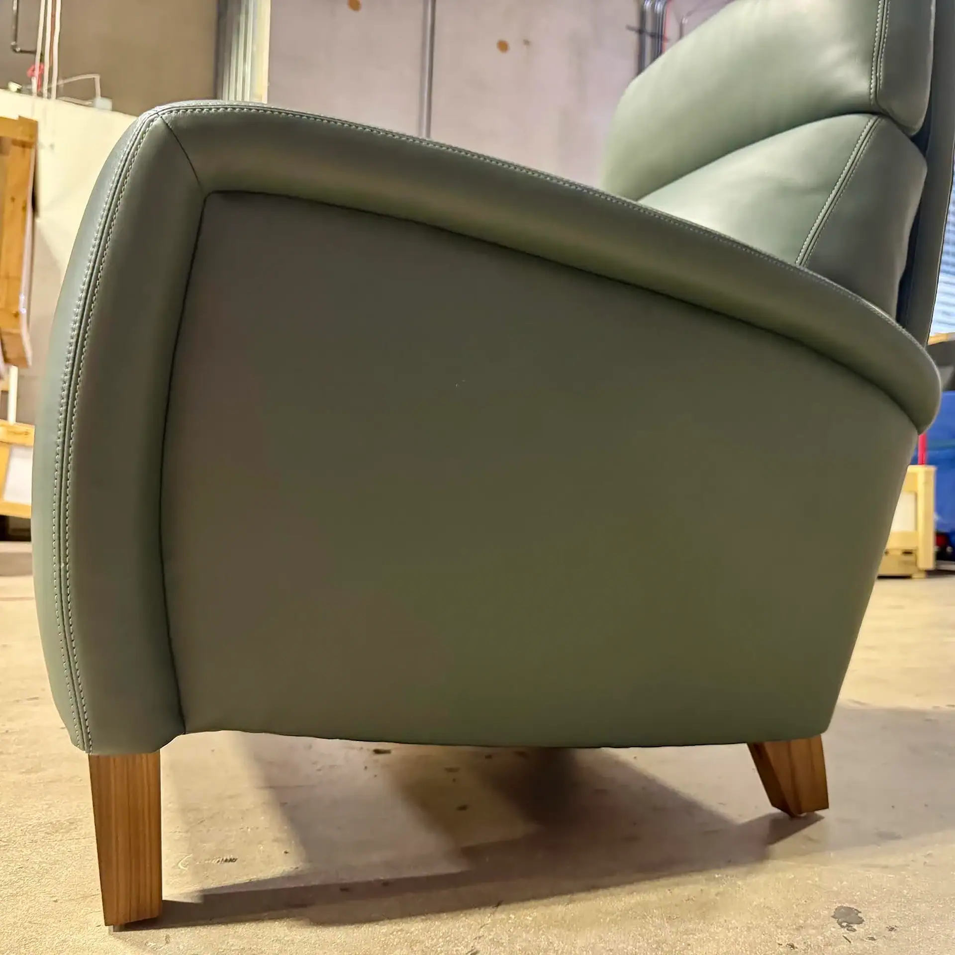 Side view of a sage green upholstered armchair with natural wood legs on a concrete floor.