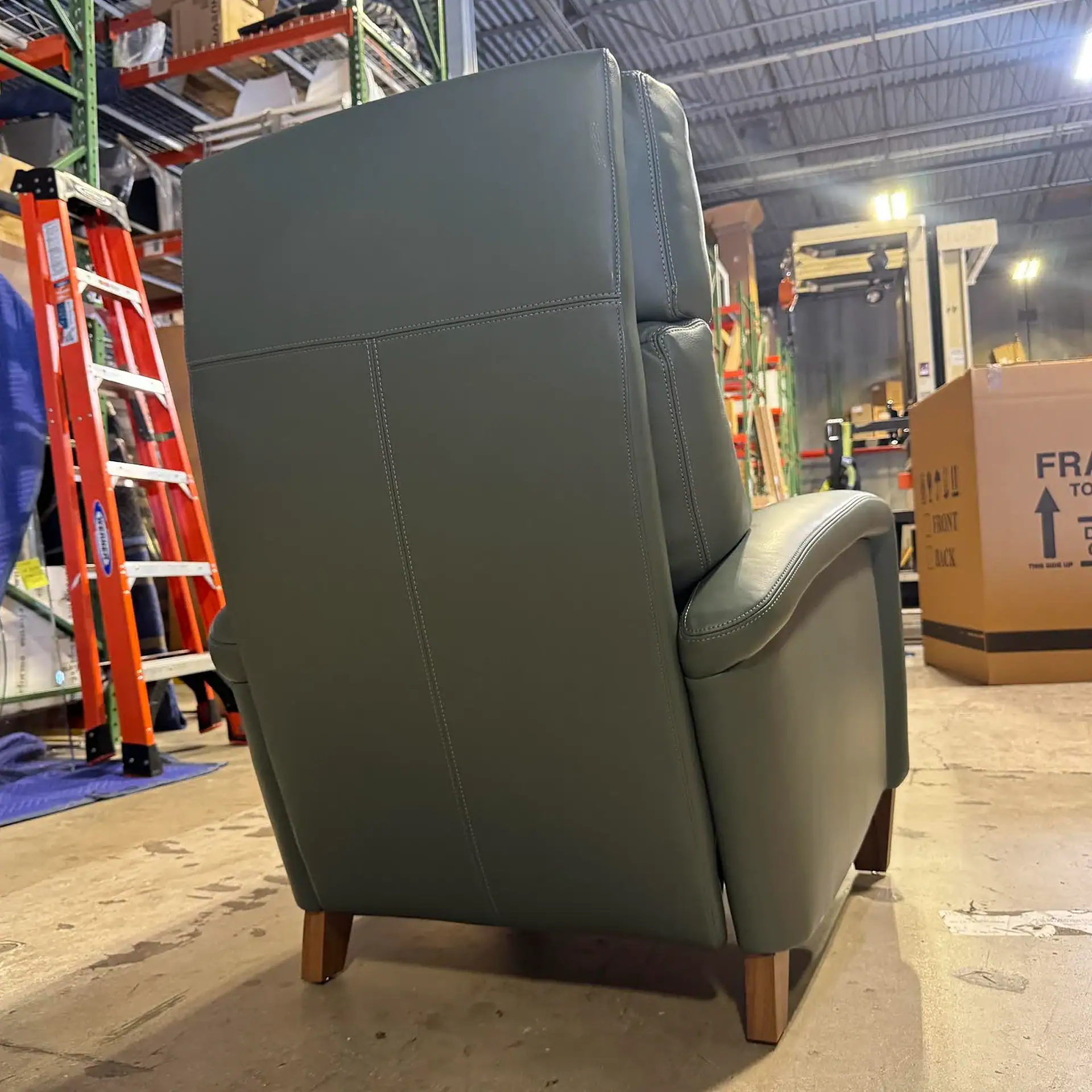 Dark green upholstered chair with wooden legs, viewed from the back-right in a warehouse.