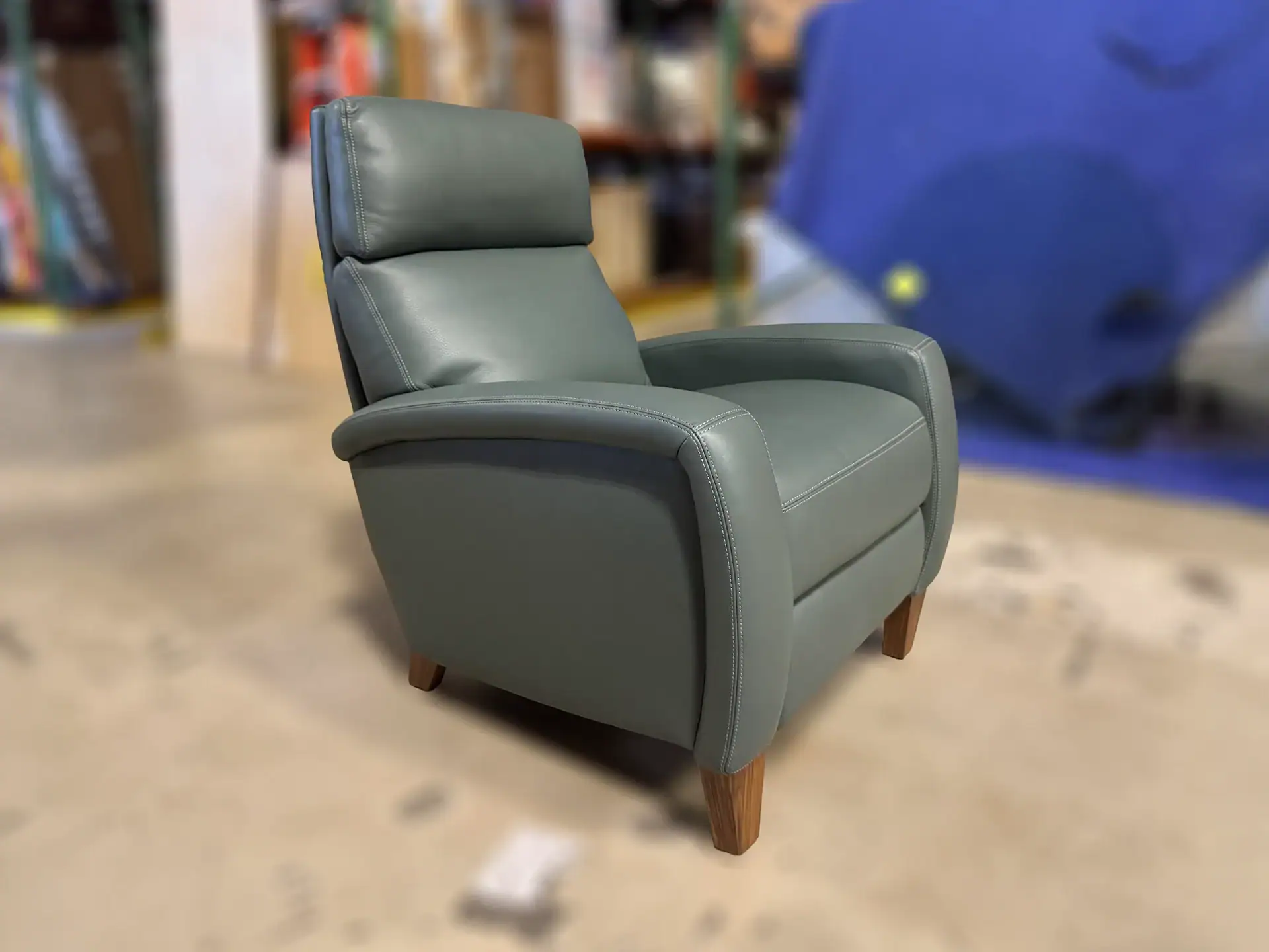 A sage green leather recliner chair with contrast stitching and wooden legs, set against a blurred indoor background.