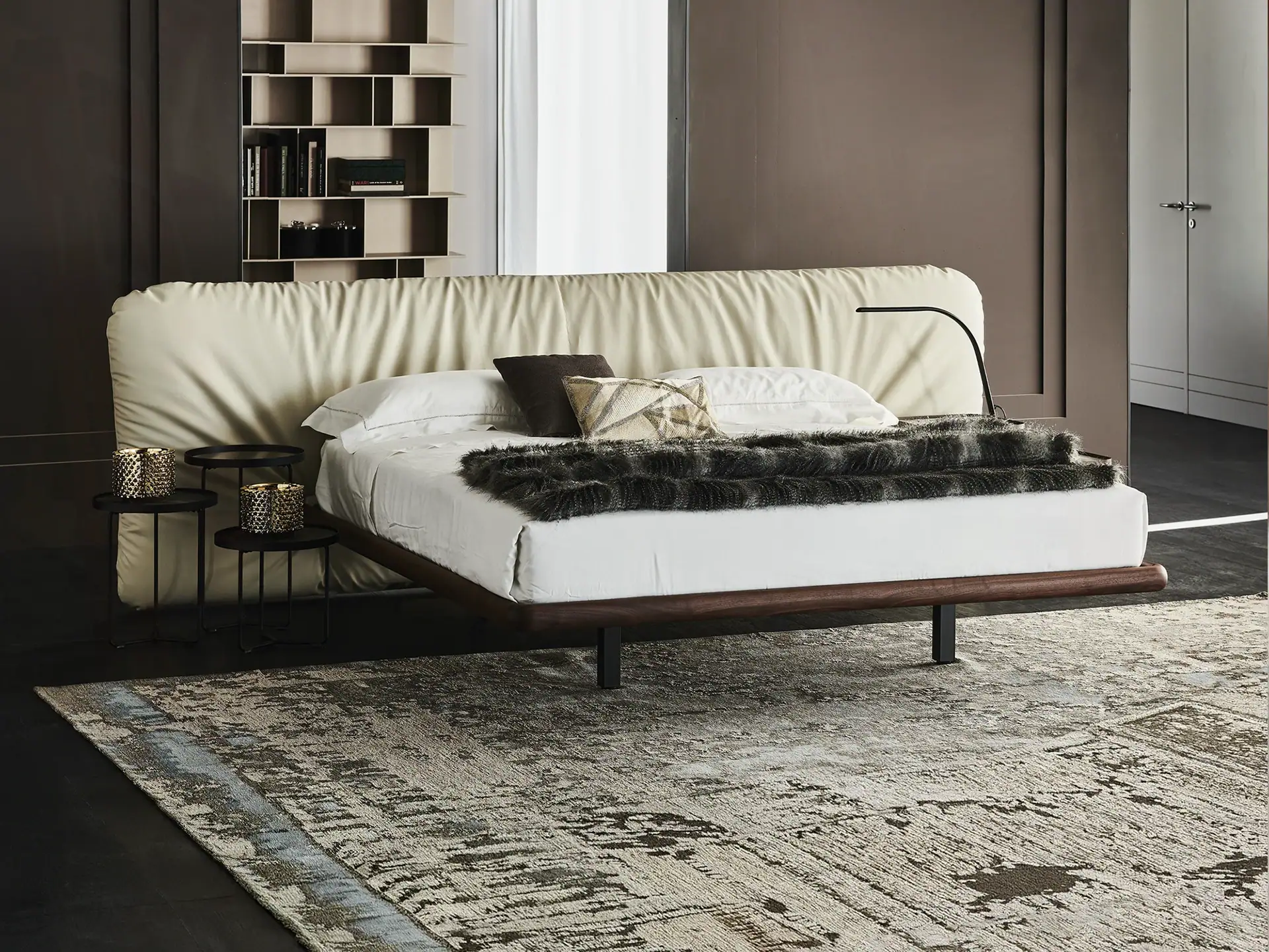 A modern bedroom with a cream upholstered bed, dark wood frame, white bedding, and a faux fur throw. Two black side tables with gold decor are on the left, and a black lamp is on the right. An abstract rug covers a dark floor. A shelving unit and a door are visible in the background.