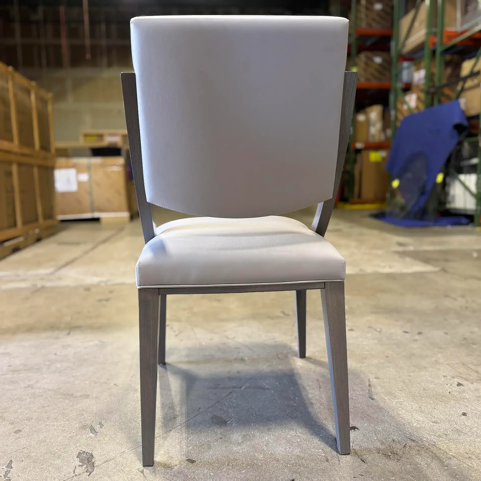 A light gray upholstered chair with a wooden frame, viewed from the back, in a warehouse setting.