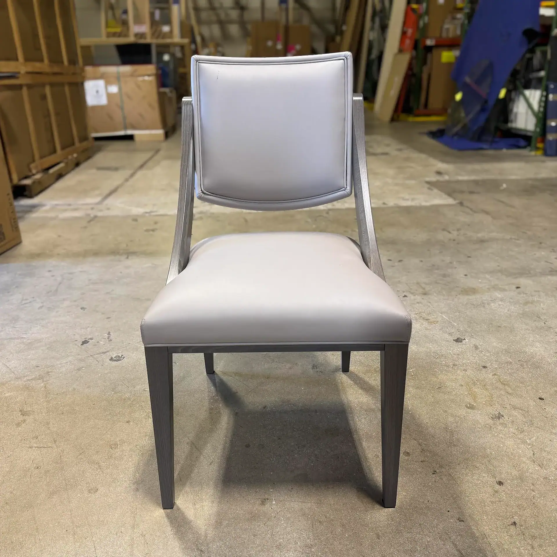 A light gray upholstered dining chair with a gray wooden frame sits on a concrete floor in a warehouse.