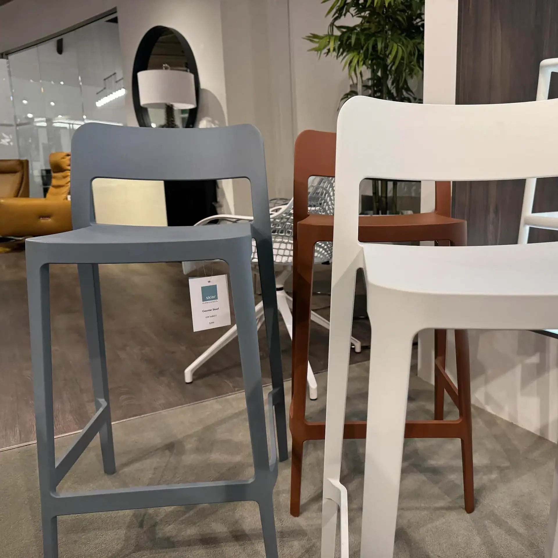 Three modern counter stools in grey, brown, and white in a showroom. The grey stool has a price tag of $399.
