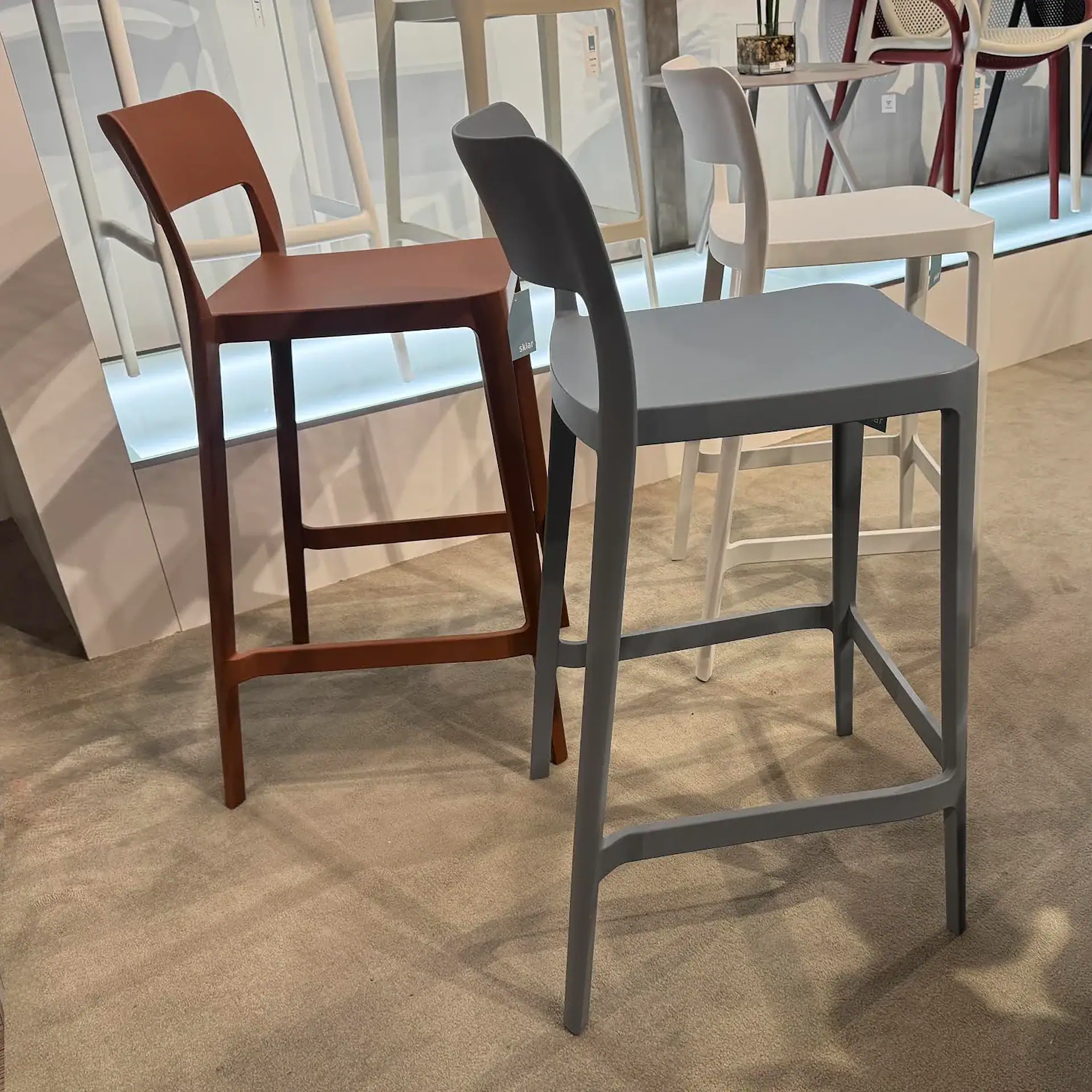 Two modern bar stools, one terracotta and one grey, stand on a beige carpet in a showroom. Other chairs and a small table are visible in the background.