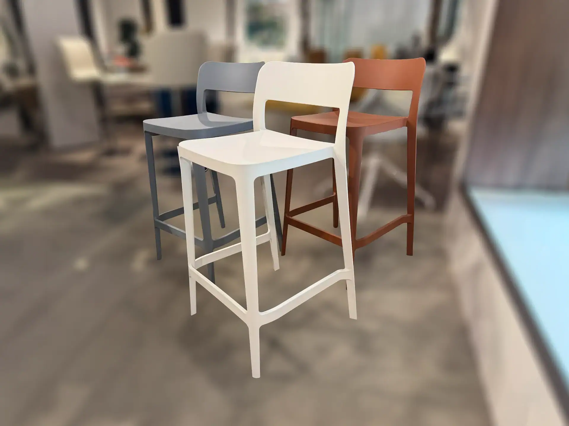Three modern bar stools in gray, off-white, and terracotta orange, arranged on a concrete floor with a blurred background.