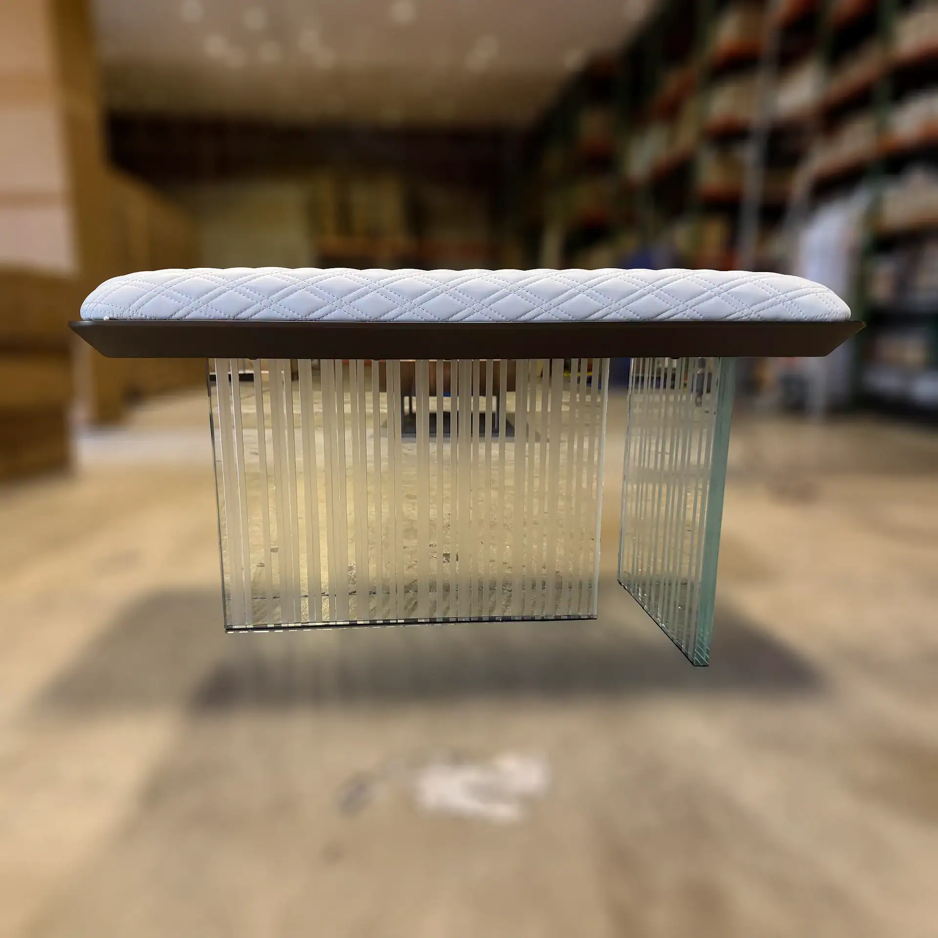 A modern bench with a white quilted cushion and transparent glass legs with vertical frosted strips, set against a blurred indoor background.