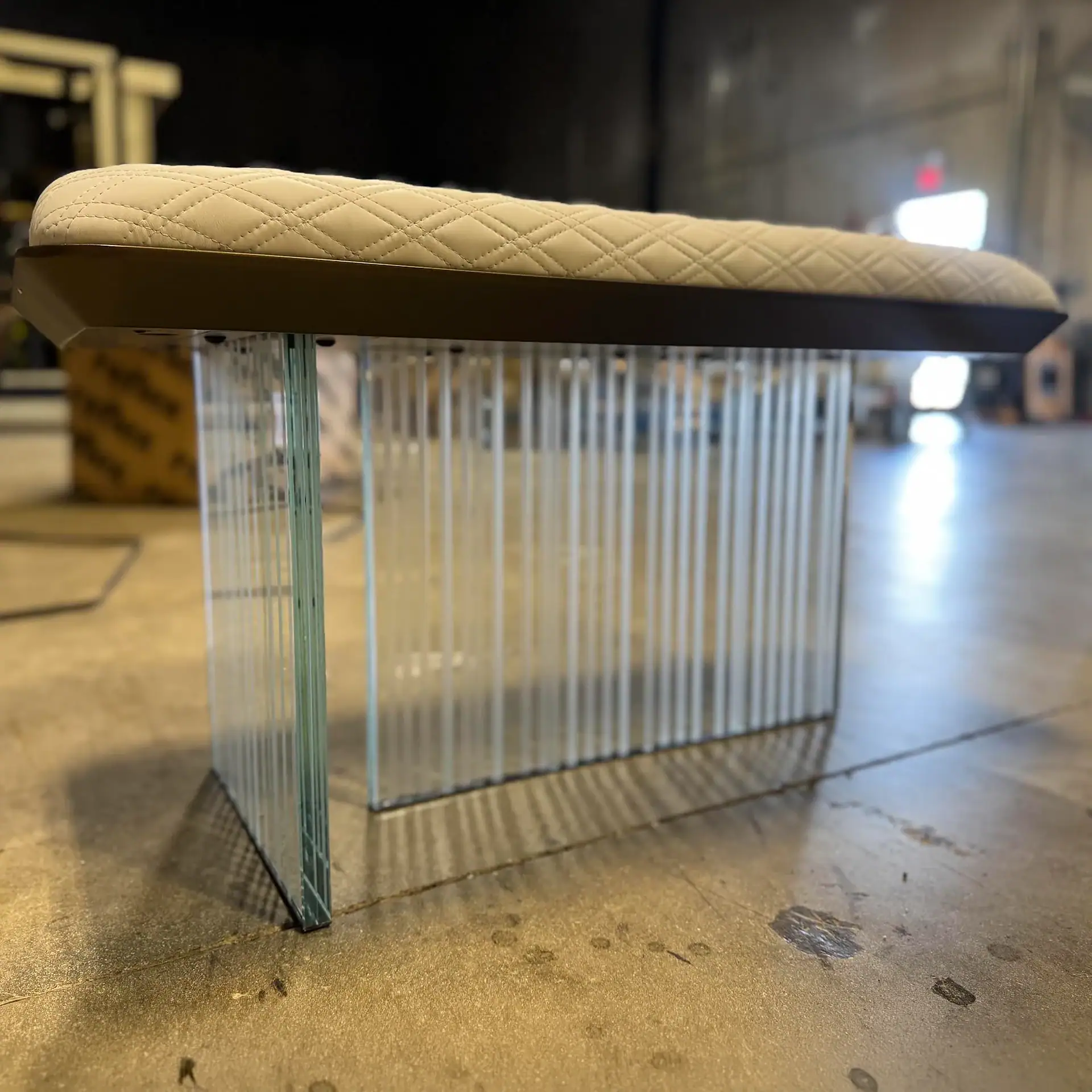 A modern bench with a cream quilted upholstered top and a transparent base made of vertical glass panels, set on a concrete floor.