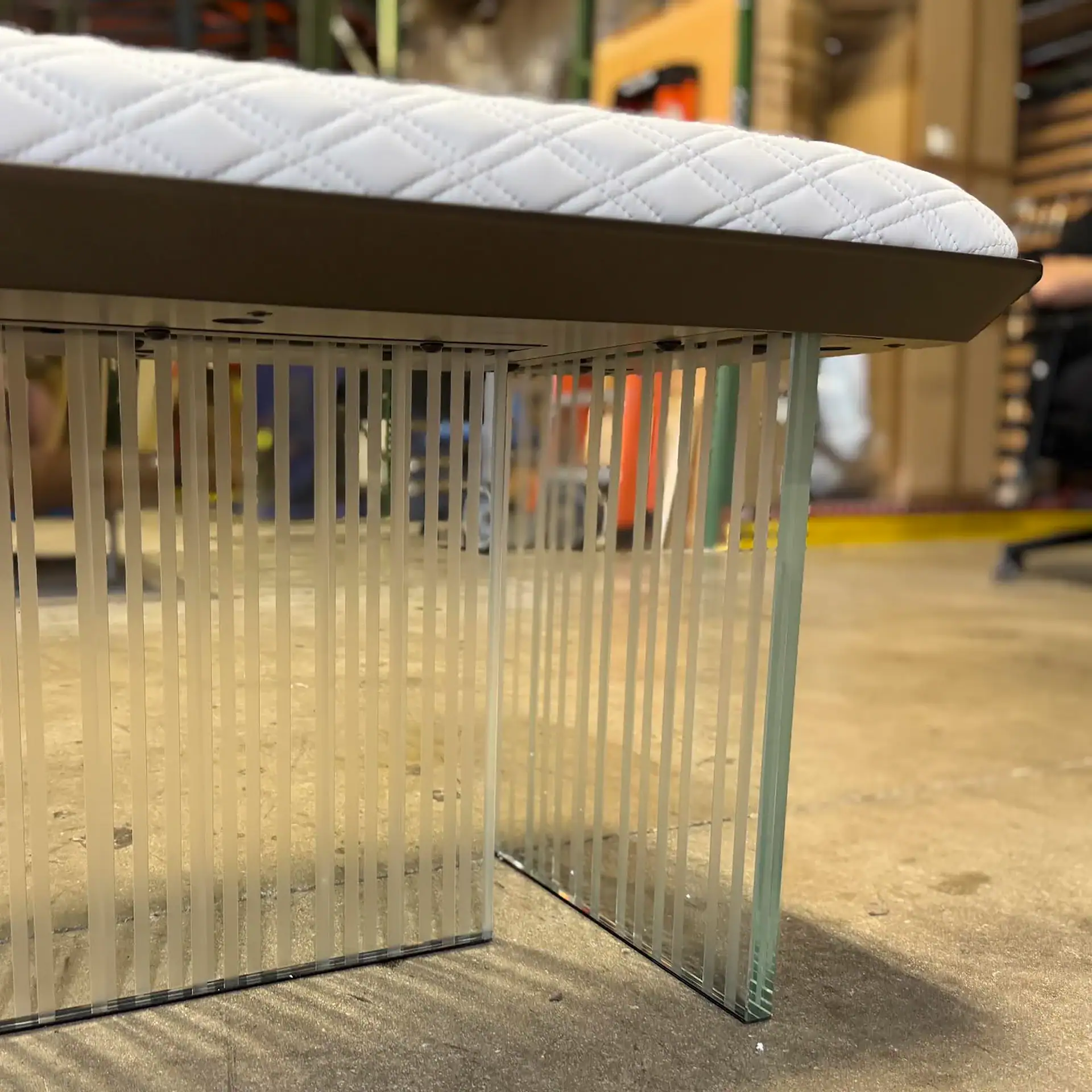 A white quilted bench with a diamond pattern top and a base made of vertical translucent strips and clear glass panels, set on an industrial floor.