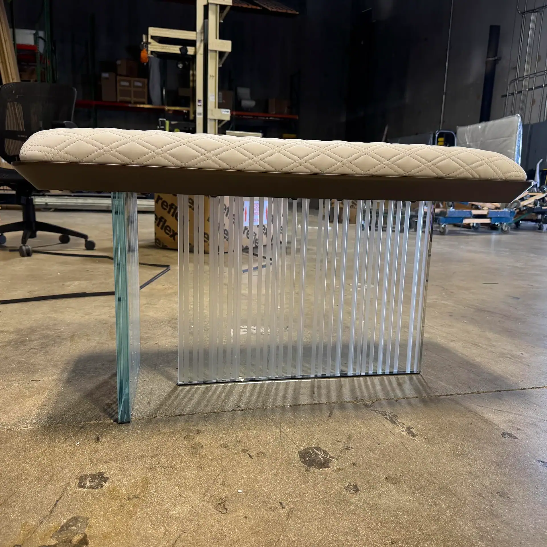 A modern bench with a quilted light-colored top and a base made of clear and frosted glass panels, set on a concrete floor in an industrial background.