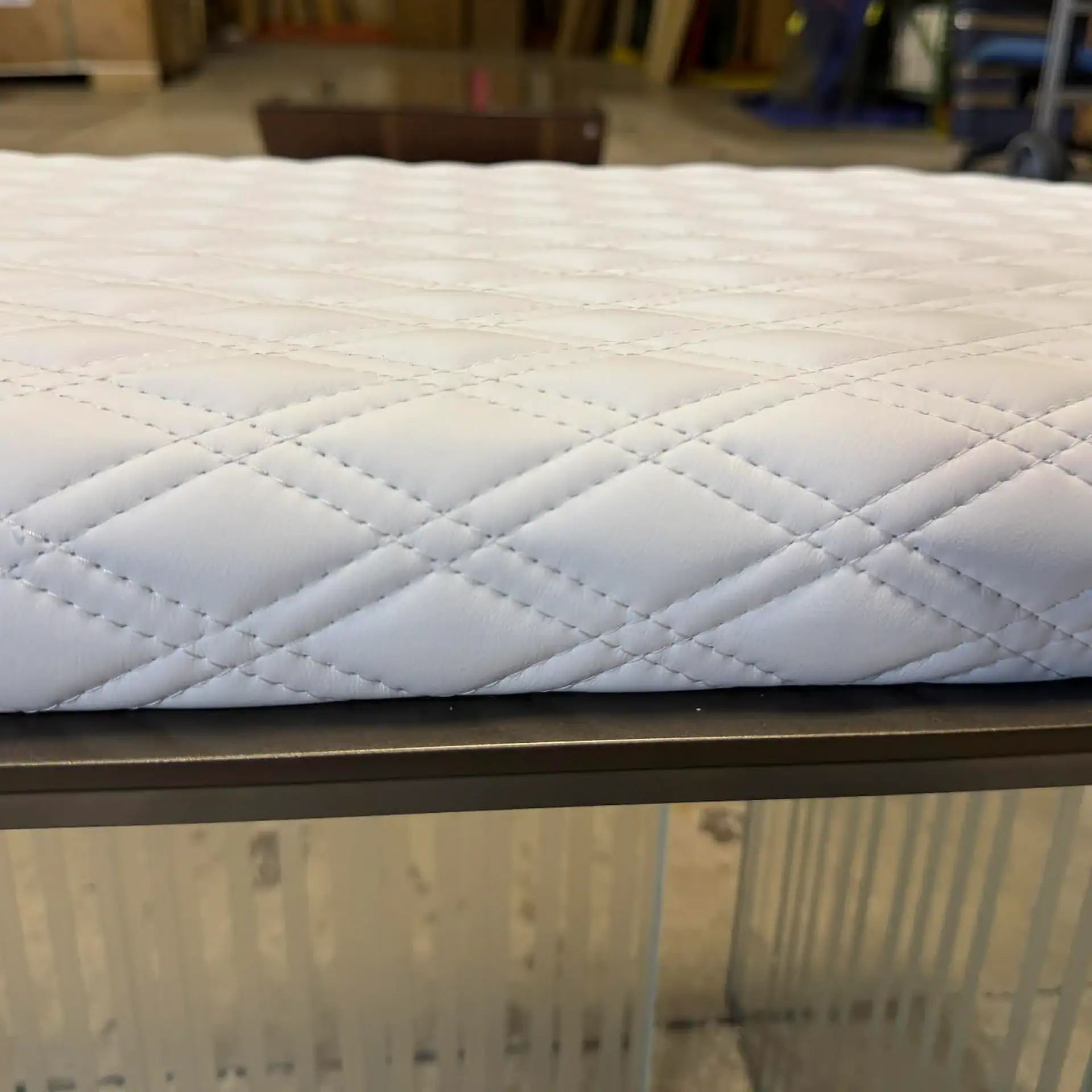 A close-up of a white quilted surface with diamond stitching, resting on a dark frame above a reflective base.