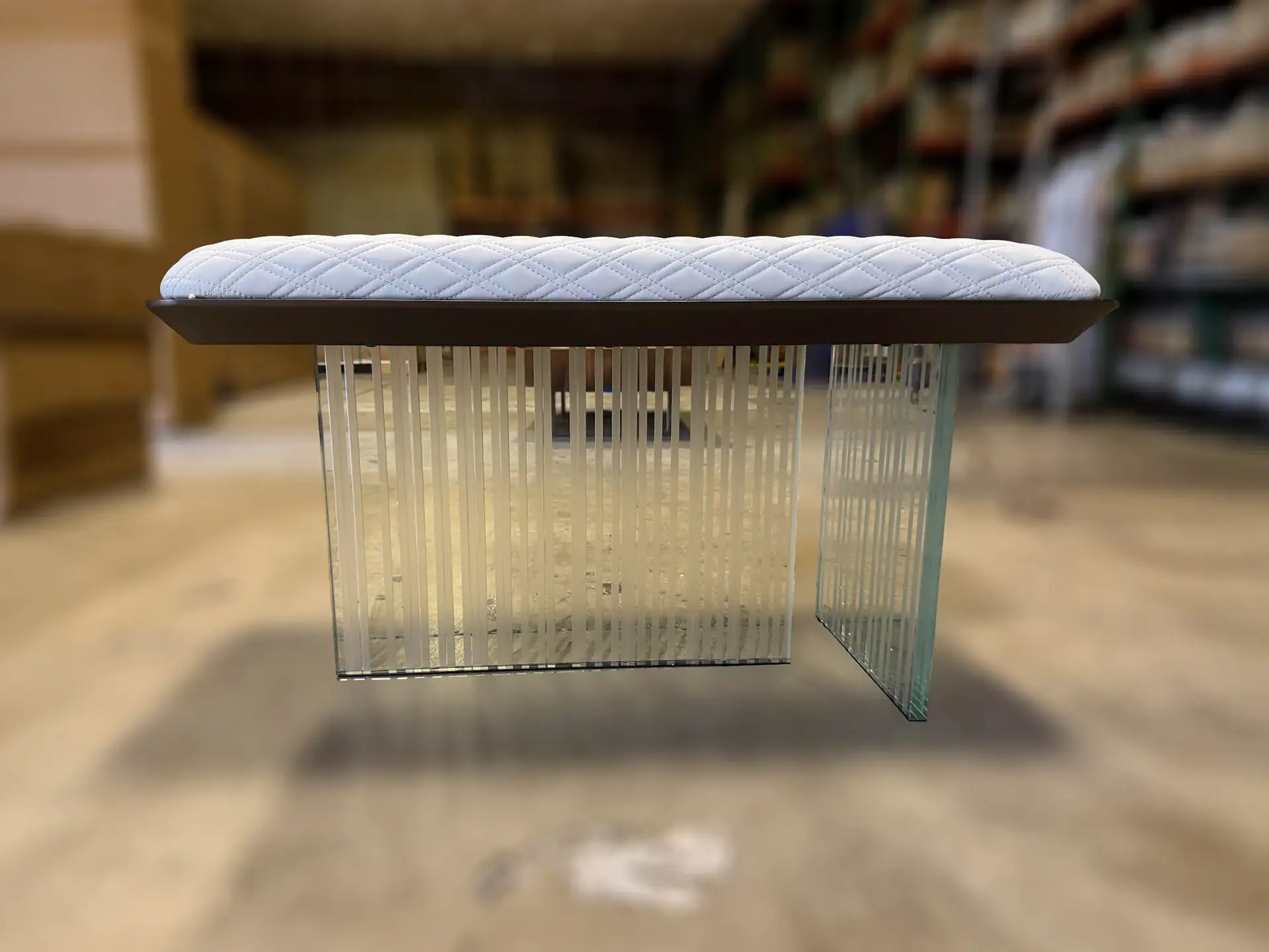A modern bench with a white quilted cushion and a clear glass base featuring frosted vertical lines, set against a blurry warehouse background.