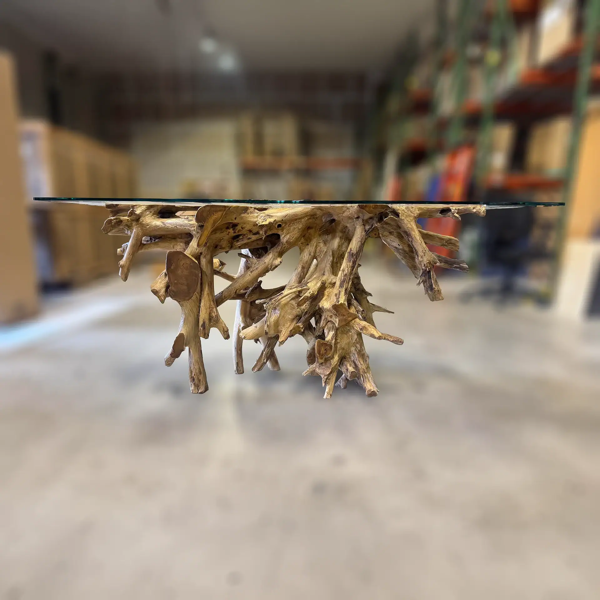 A glass-top table with a gnarled natural wood root base, set against a blurred warehouse background.