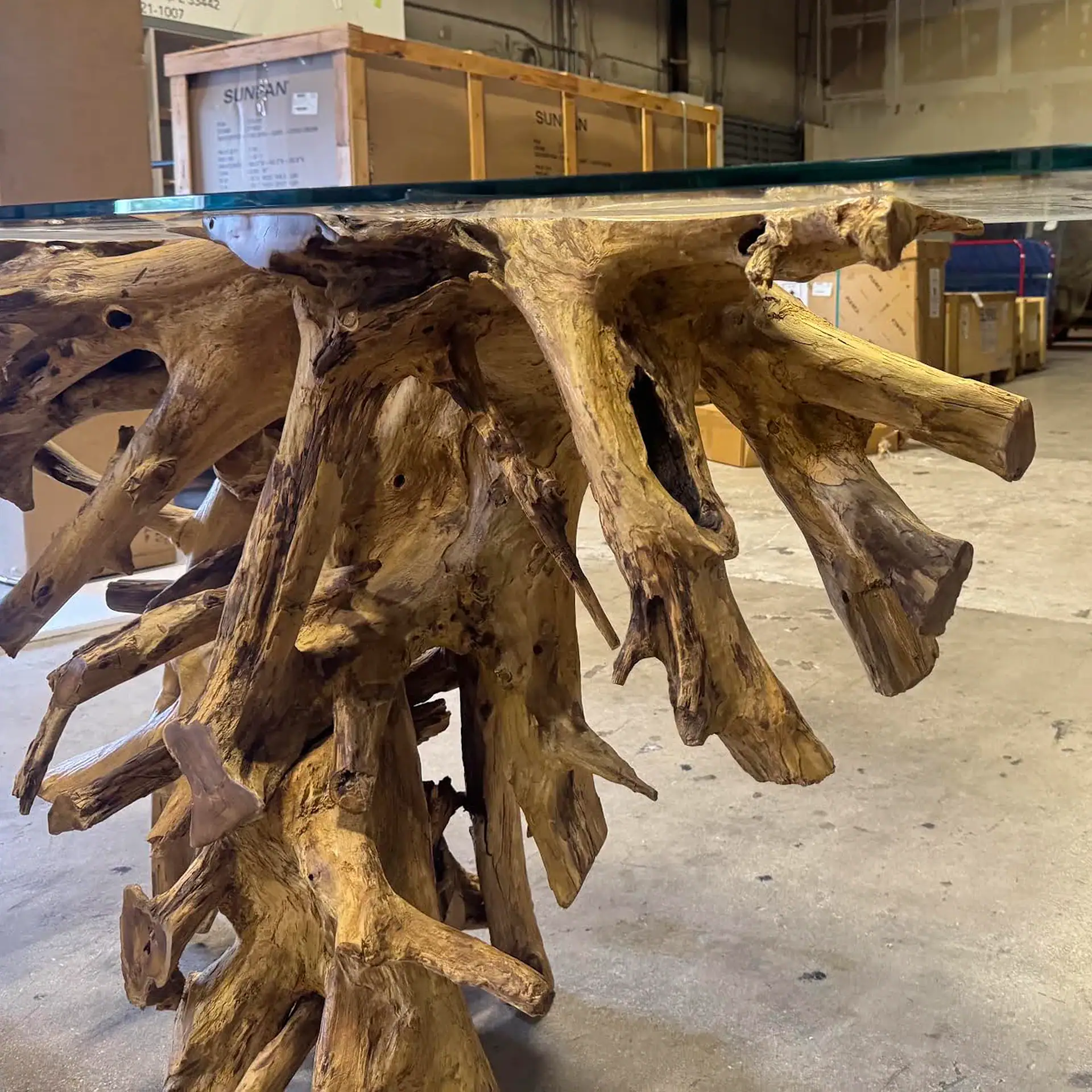 A table with a clear glass top and a base made of natural, gnarled wood roots, set in a warehouse.