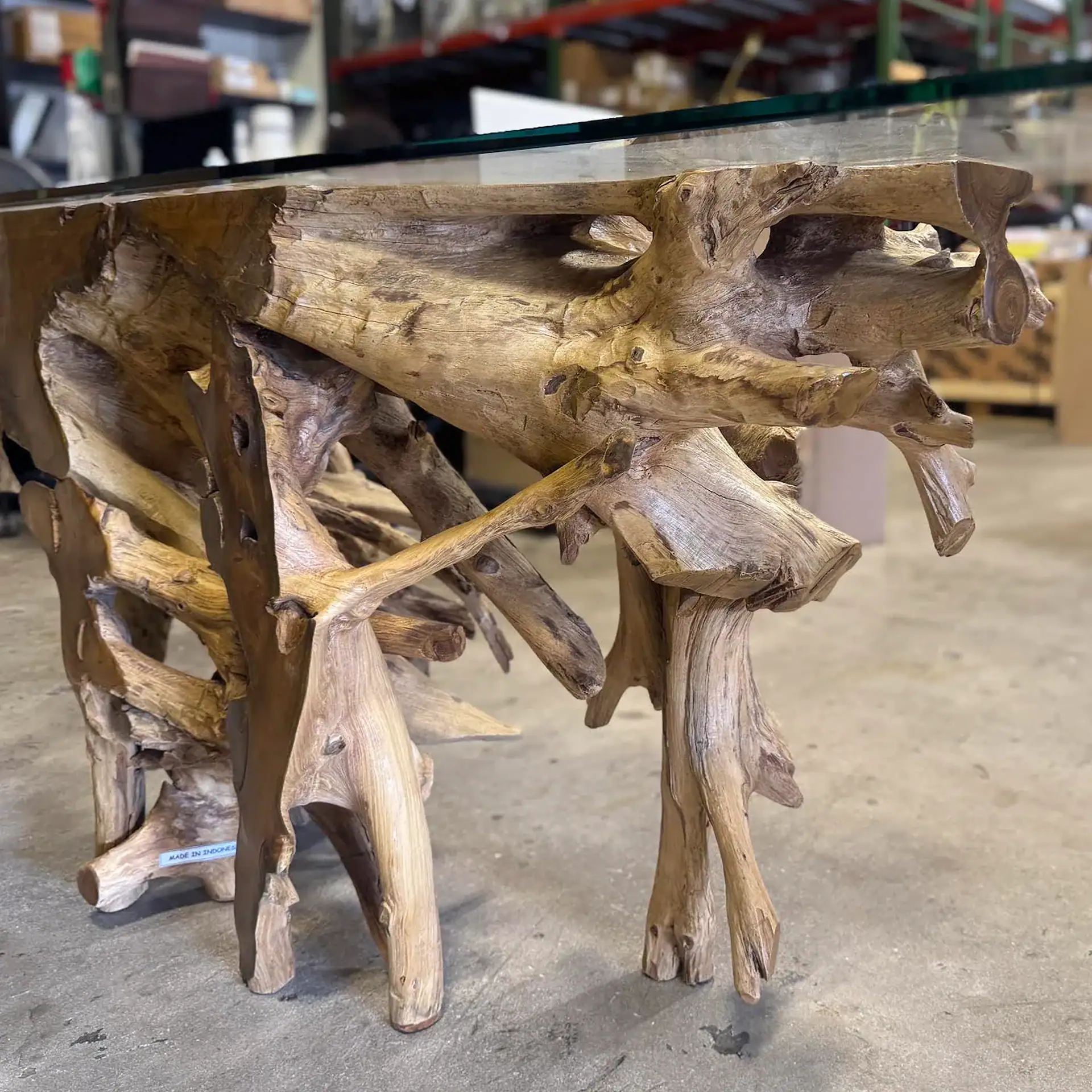 A console table with a clear glass top and a base made from natural, light brown, intricately shaped wood roots. A label on the base reads 'MADE IN INDONESIA'.