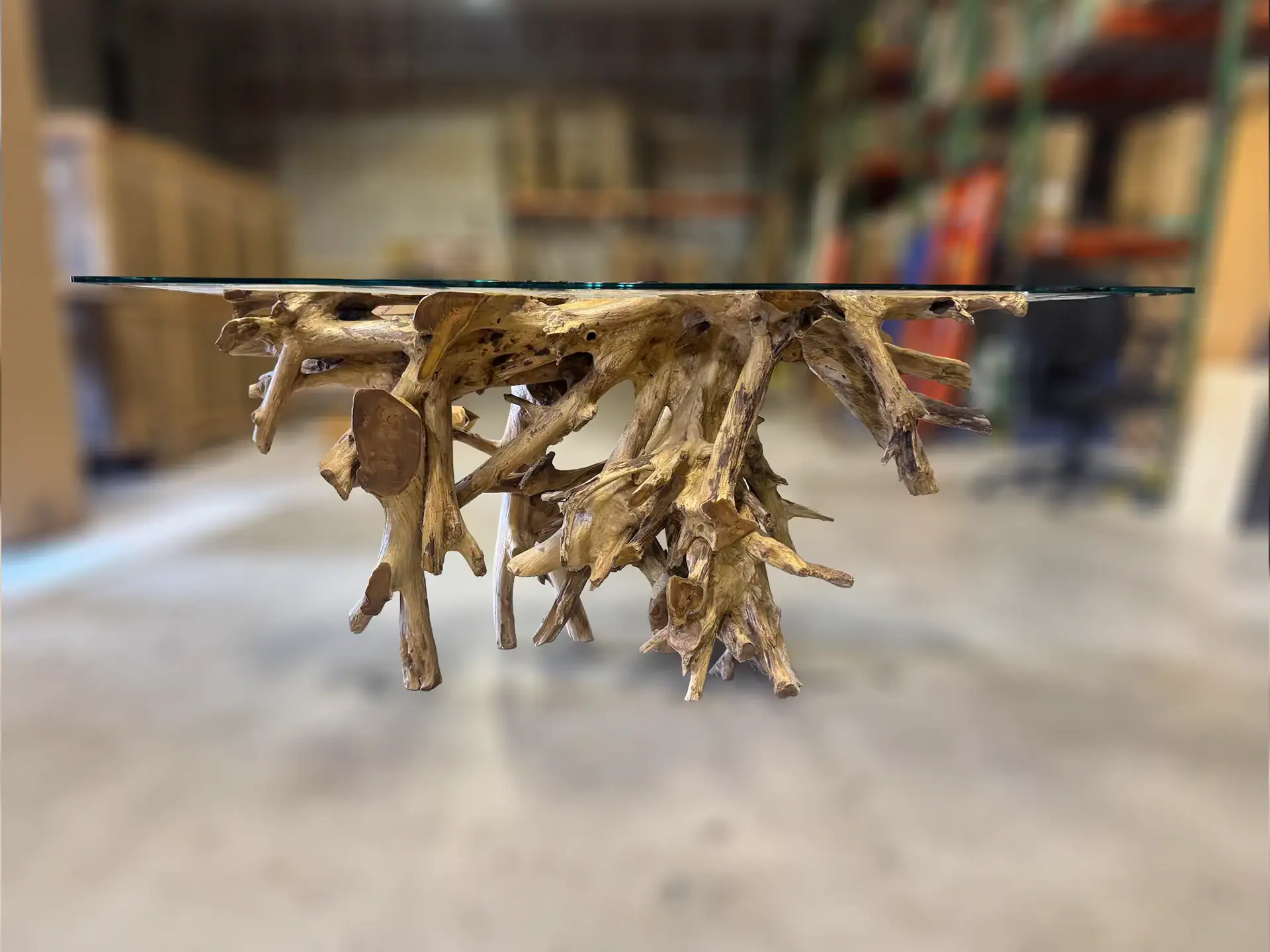 A glass-top table with a gnarled, natural wood root base, set against a blurred warehouse background.