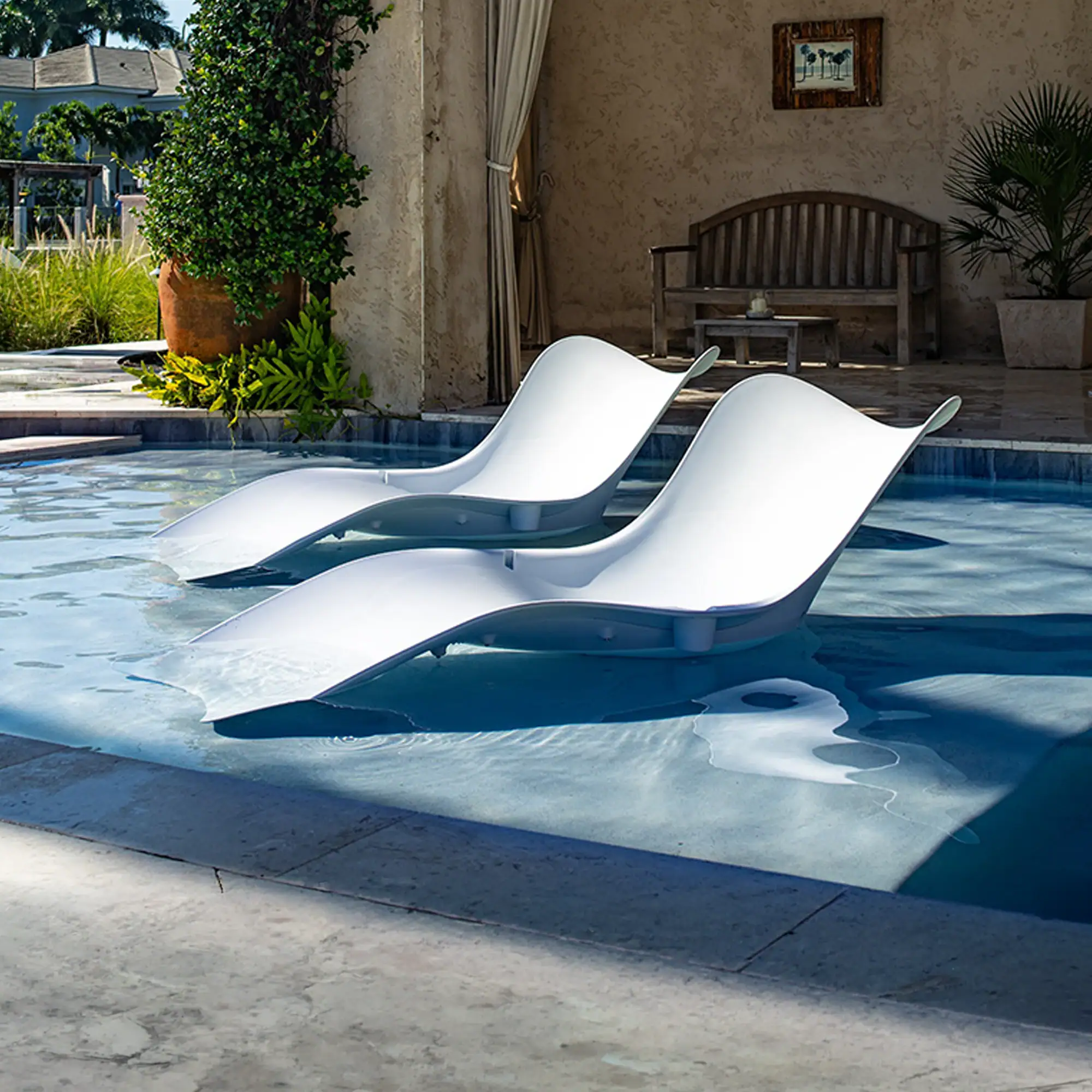 Two white lounge chairs partially submerged in a swimming pool, with a patio and lush greenery in the background.