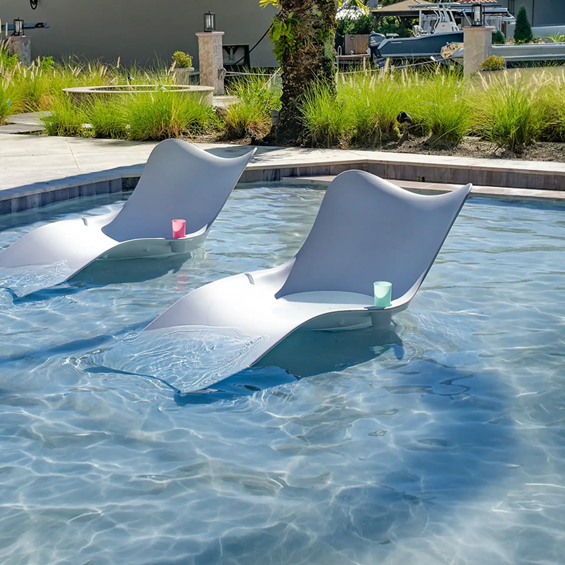 Two light gray, modern lounge chairs partially submerged in a clear blue swimming pool. One chair holds a pink cup, the other a light green cup. Green grasses and a concrete patio surround the pool under a bright sky.