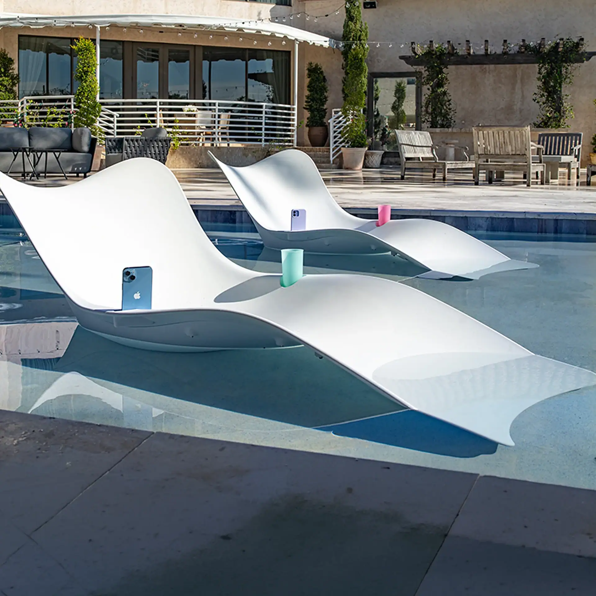 Two white, modern lounge chairs are partially submerged in a clear blue swimming pool, each holding a smartphone and a cup. A luxurious patio with outdoor furniture and a building with glass doors is in the background.