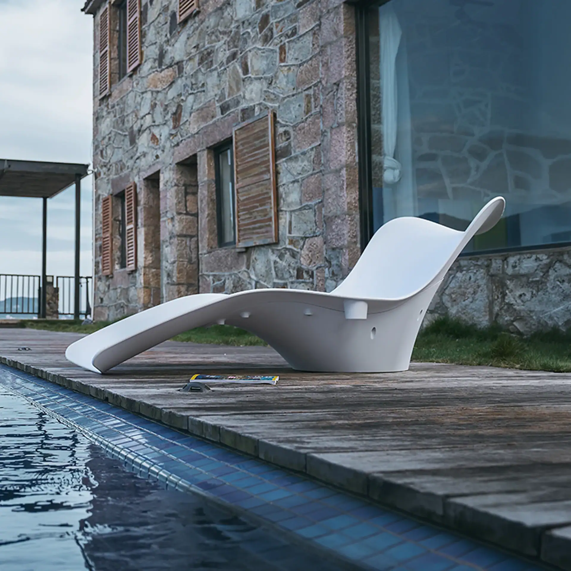 A white, S-shaped lounge chair on a wooden deck next to a swimming pool, with a stone building in the background.
