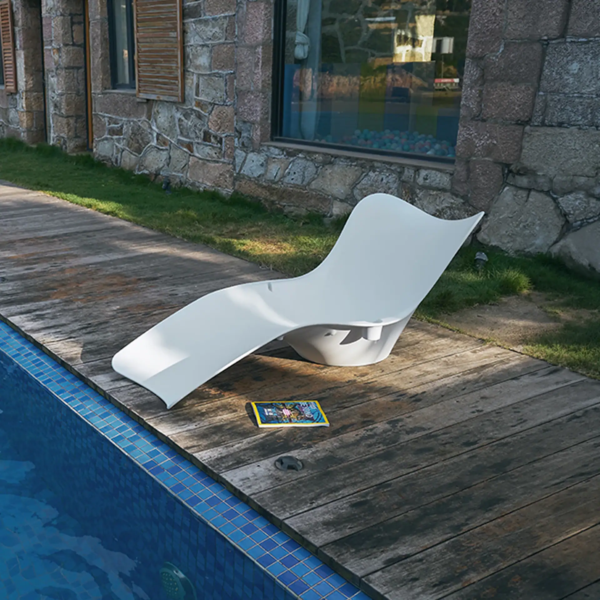 A white lounge chair on a wooden deck next to a blue swimming pool. A magazine lies on the deck. A stone building with windows and grass is in the background.