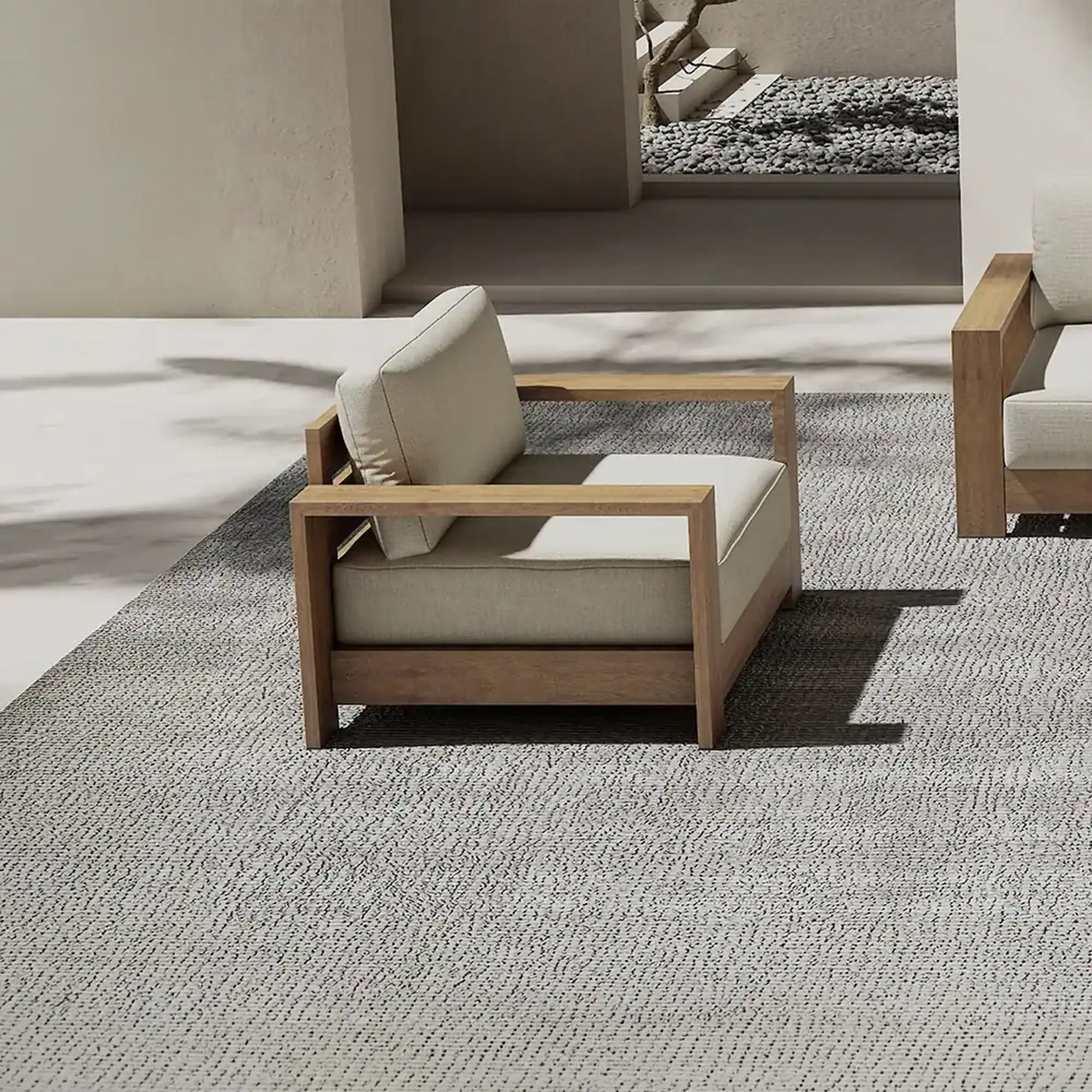 Two modern outdoor armchairs with light wooden frames and light beige cushions on a textured grey rug. A pebble garden and light walls are in the background.