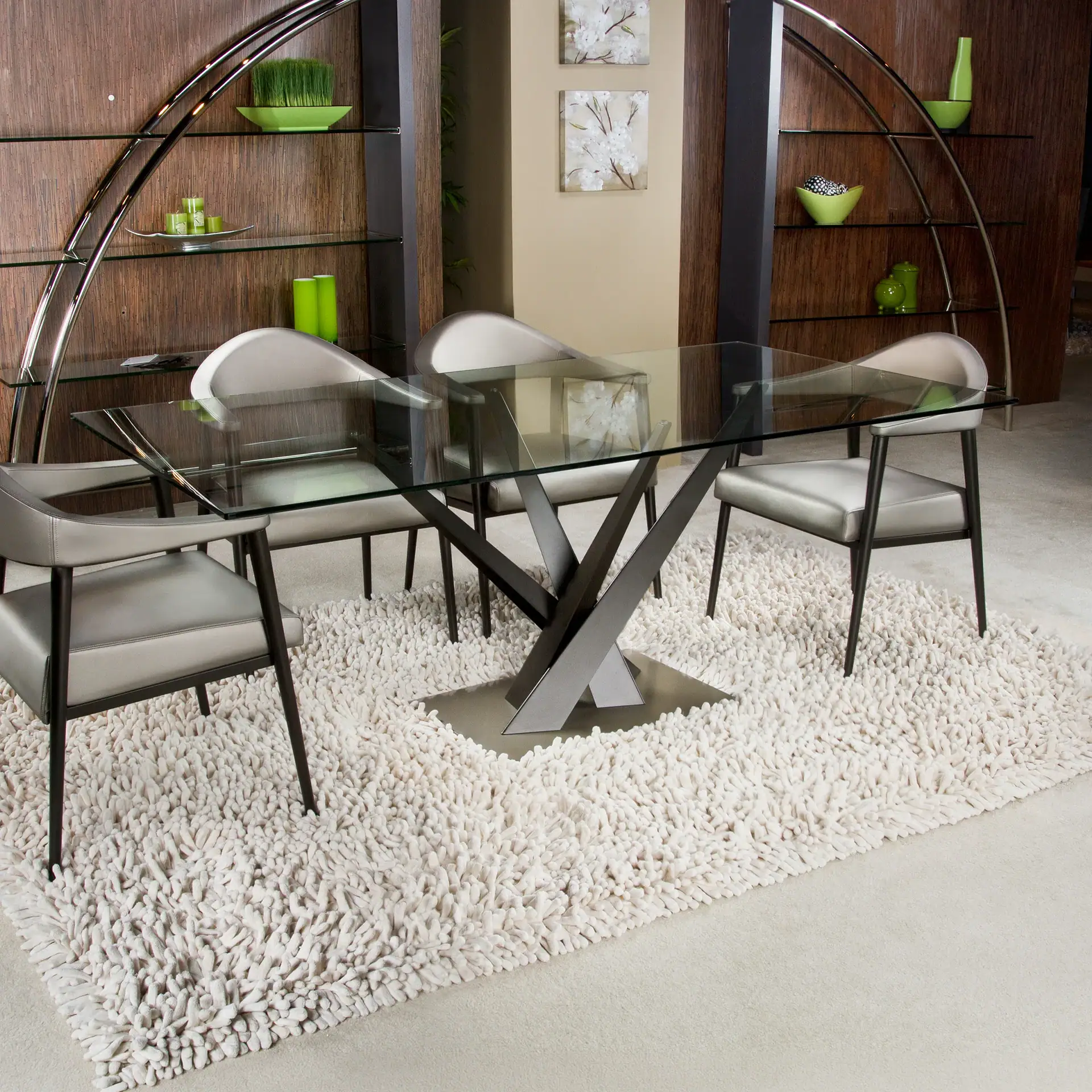 A modern dining room with a glass table, four gray chairs on a shaggy cream rug, and dark wood shelving units with green decor.