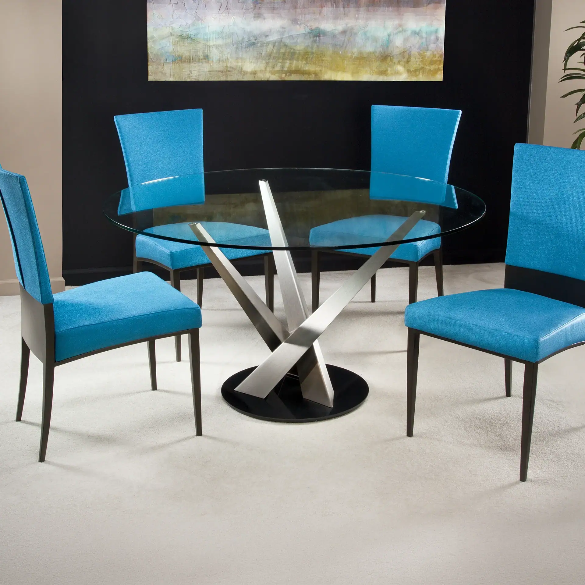 A modern dining set with a round glass table and four turquoise blue chairs on a light carpet, against a dark wall with abstract art.