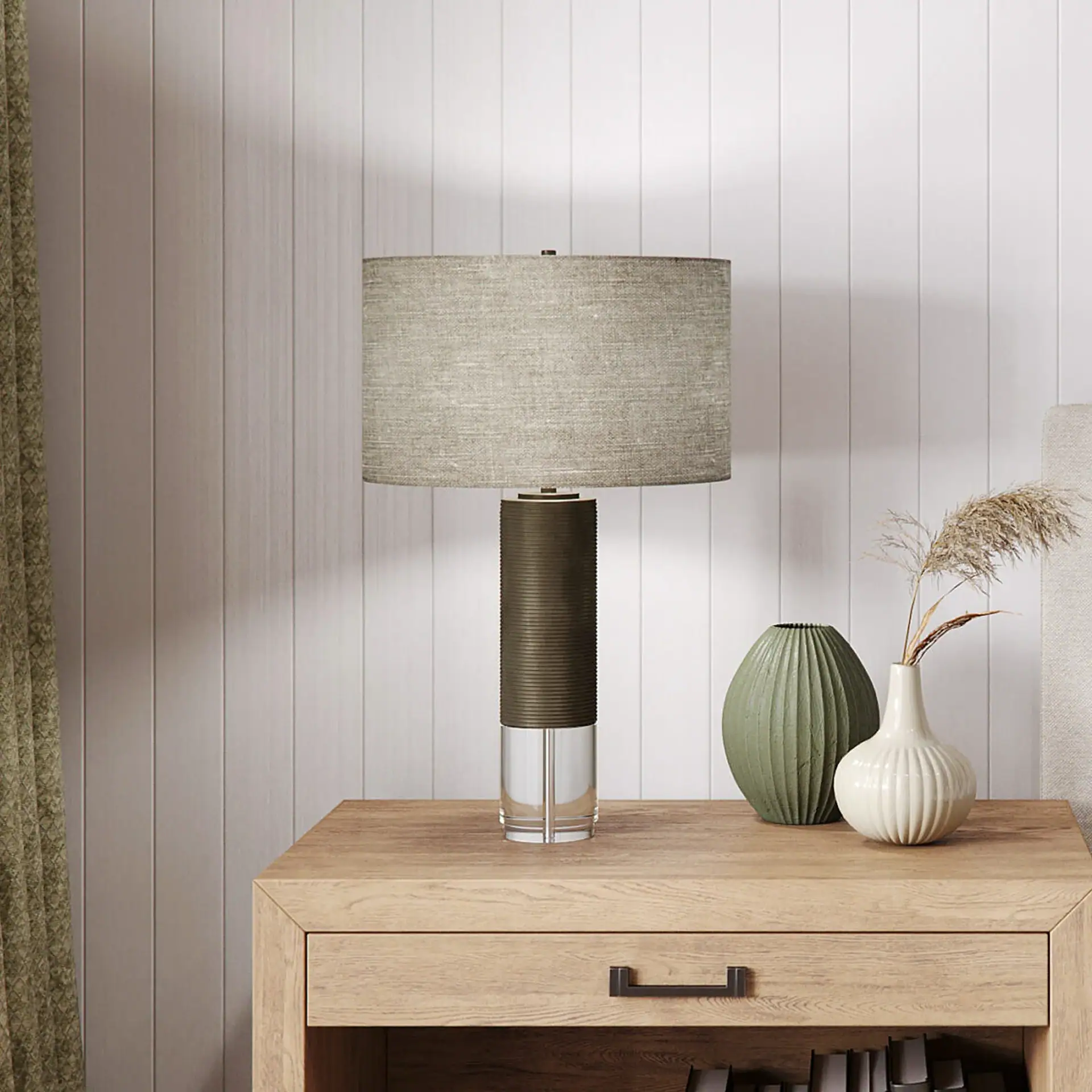 A modern table lamp with a beige shade and a dark bronze and clear base, two decorative vases with pampas grass, and a light wood nightstand with a drawer, all against a white paneled wall.