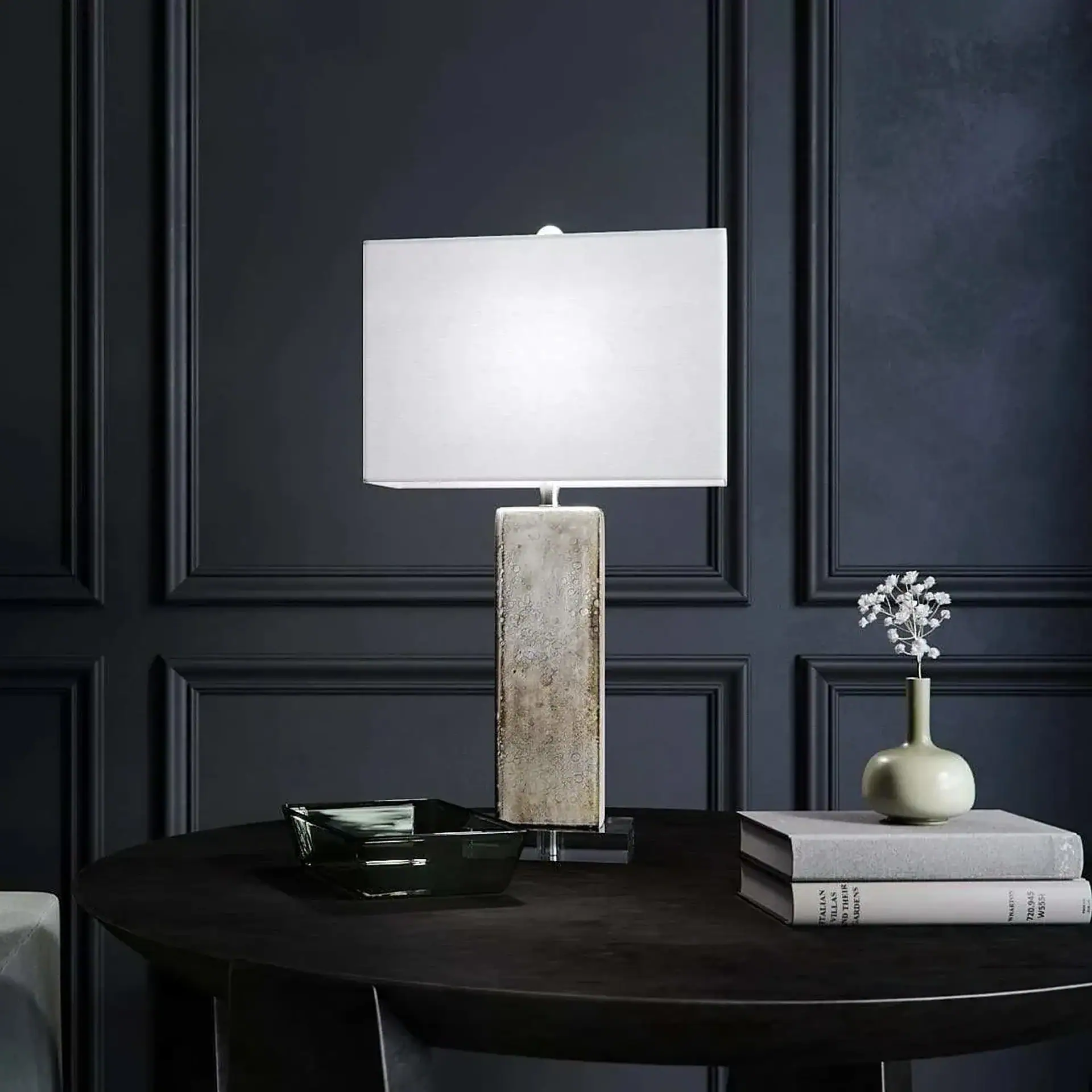 A modern table lamp with a rectangular white shade and a textured rectangular base on a dark round table. Decorative items including a glass bowl, stacked books, and a small vase with white flowers are also on the table. The background is a dark paneled wall.