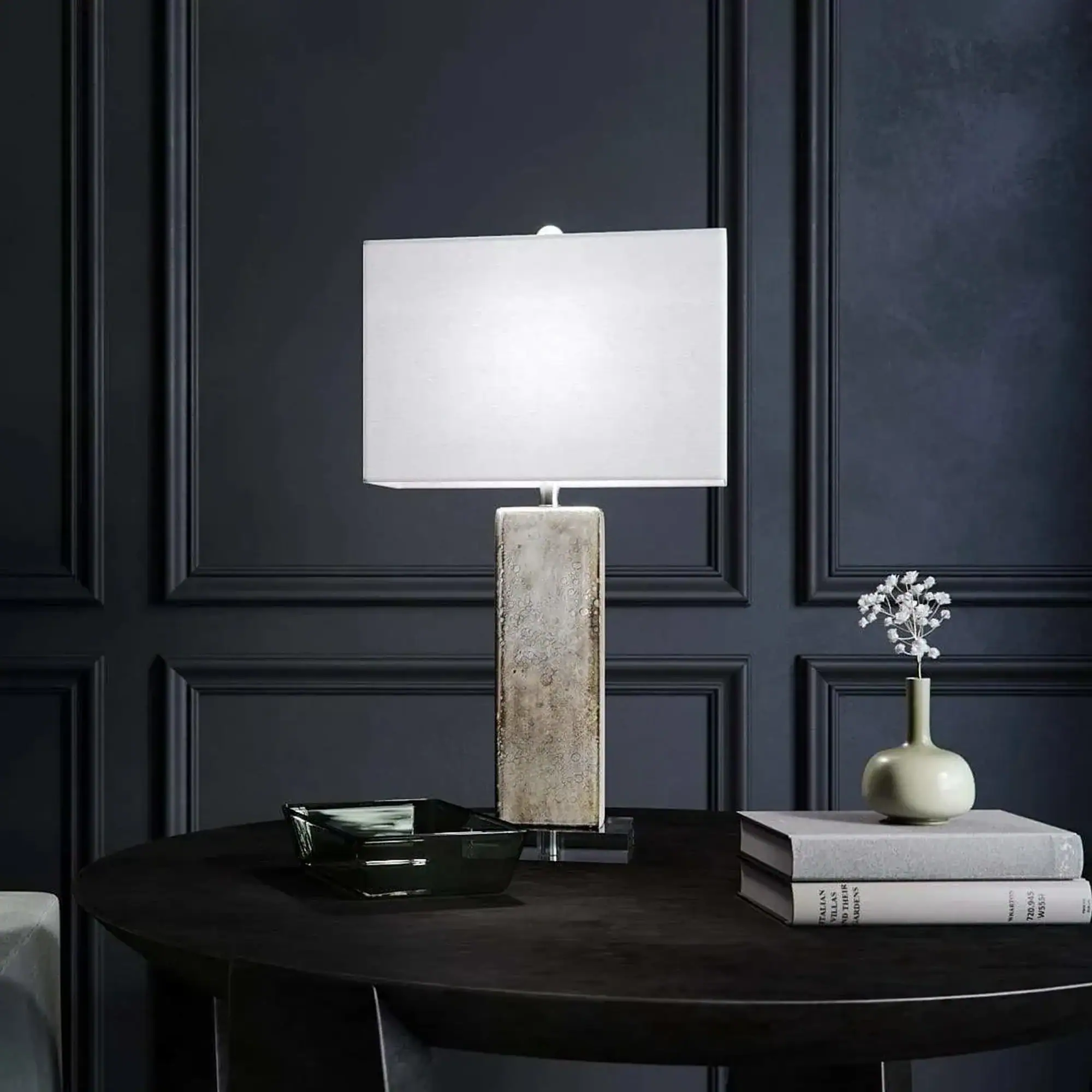 A modern table lamp with a rectangular white shade and a textured rectangular base on a dark round table. Decorative items including a glass bowl, stacked books, and a small vase with white flowers are also on the table. The background is a dark paneled wall.