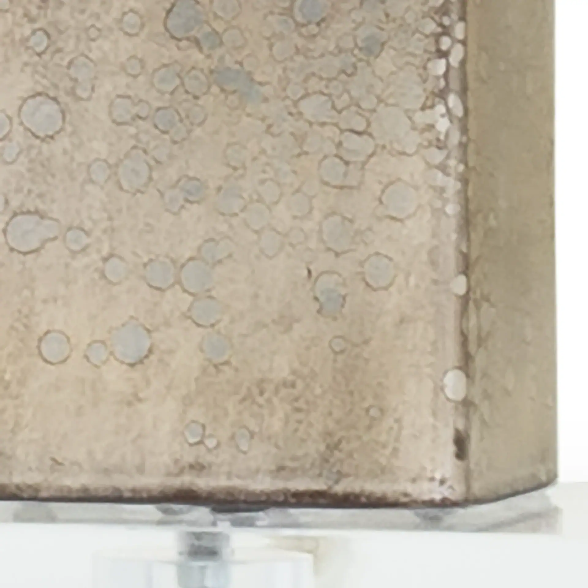 Close-up of a rectangular object with a mottled, textured metallic surface in light gray and beige, resting on a clear base.