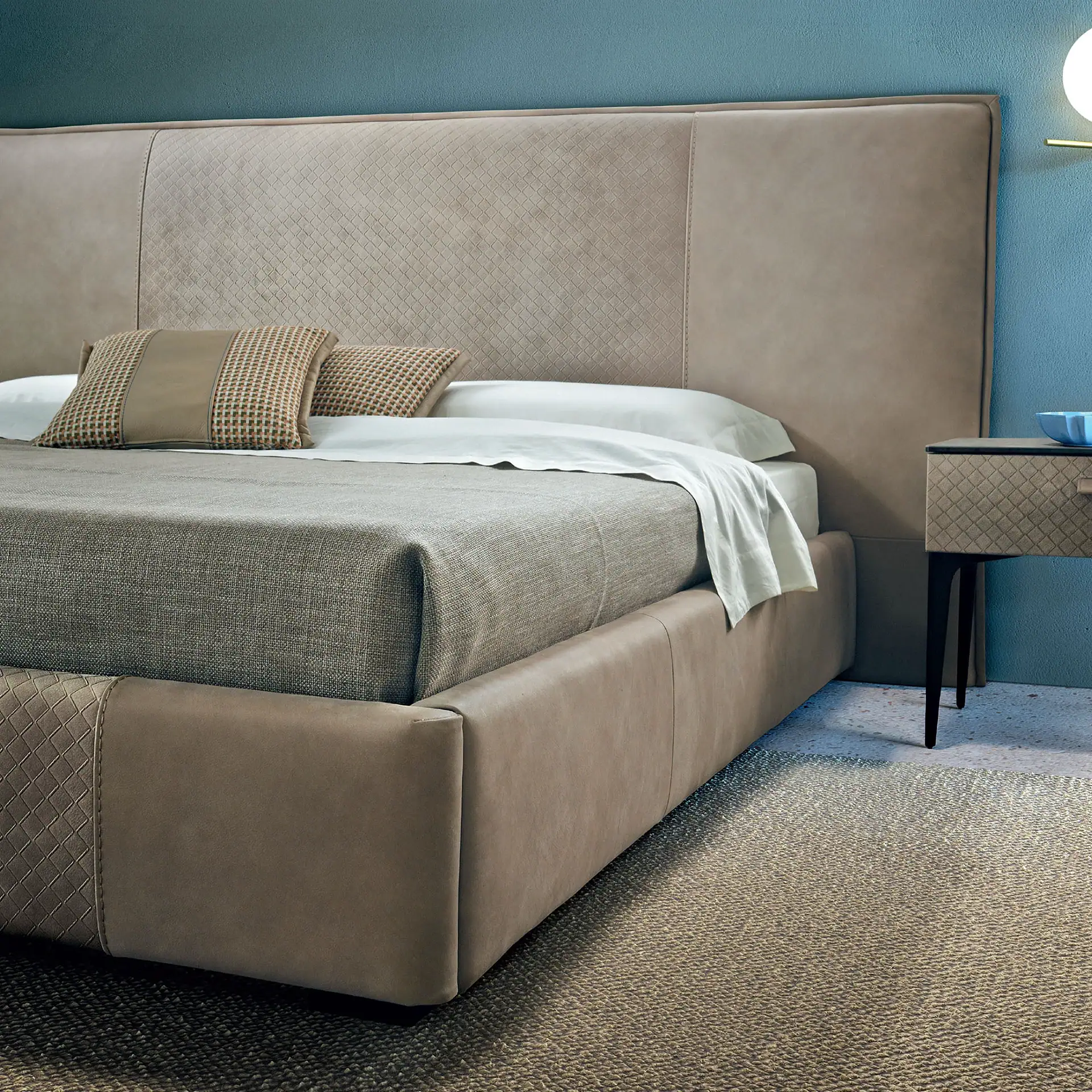 A modern bedroom with an upholstered bed, tall patterned headboard, white sheets, grey blanket, and decorative pillows. A nightstand with a blue bowl and a wall lamp are on the right against a blue-green wall, with a textured rug on the floor.