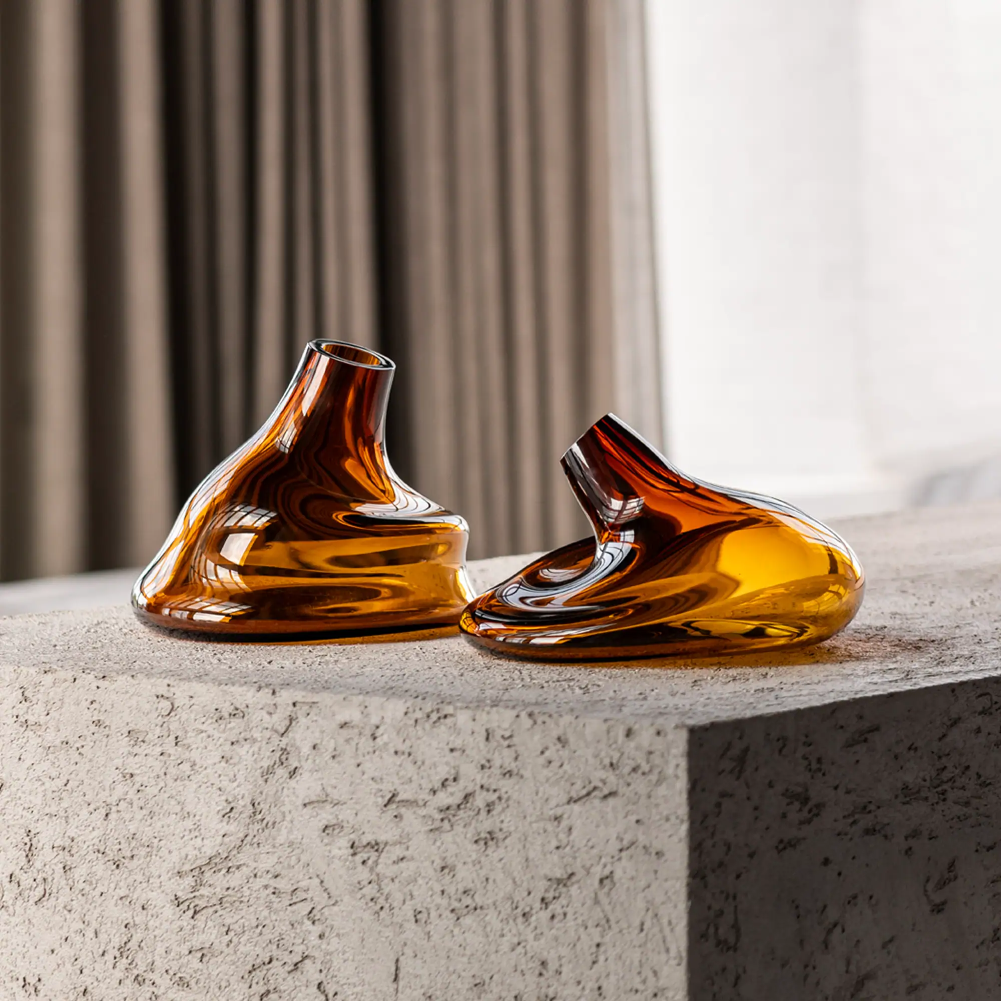 Two amber-colored, organically shaped glass vases with reflective surfaces, resting on a gray textured concrete block.