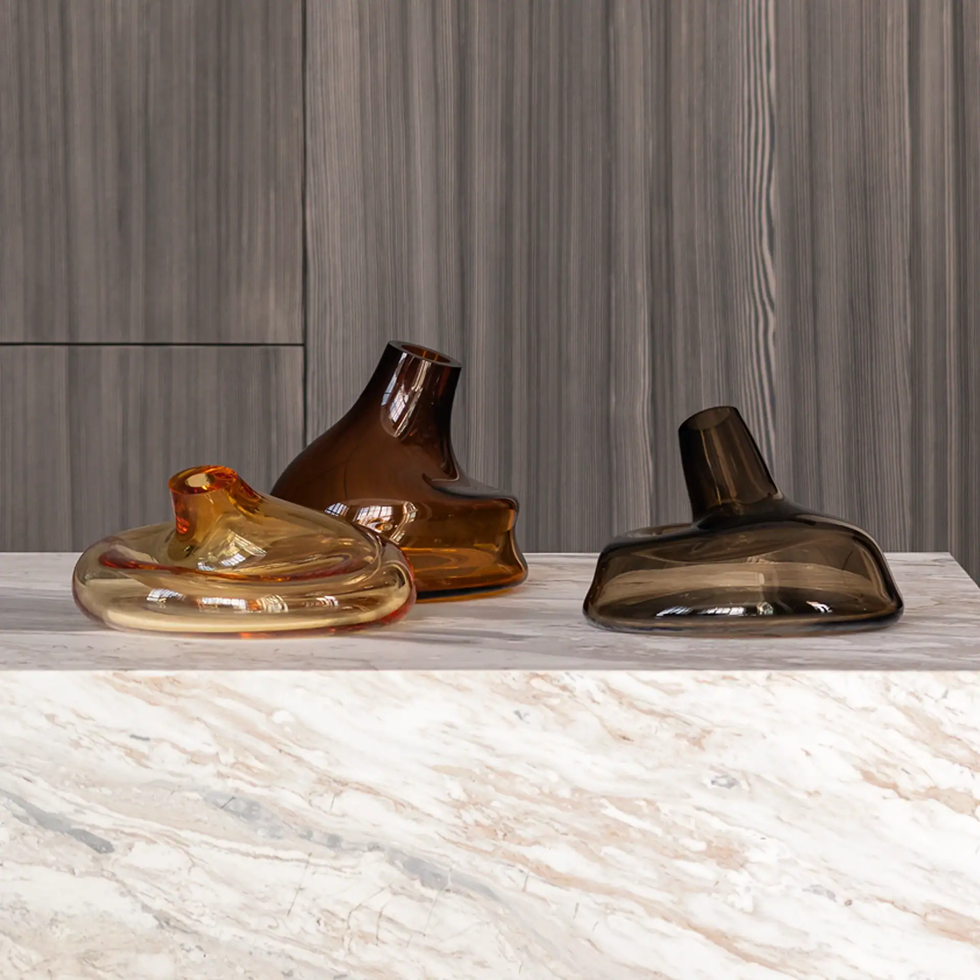 Three uniquely shaped glass decorative objects in amber and smoky brown colors on a marble surface with a dark wooden background.