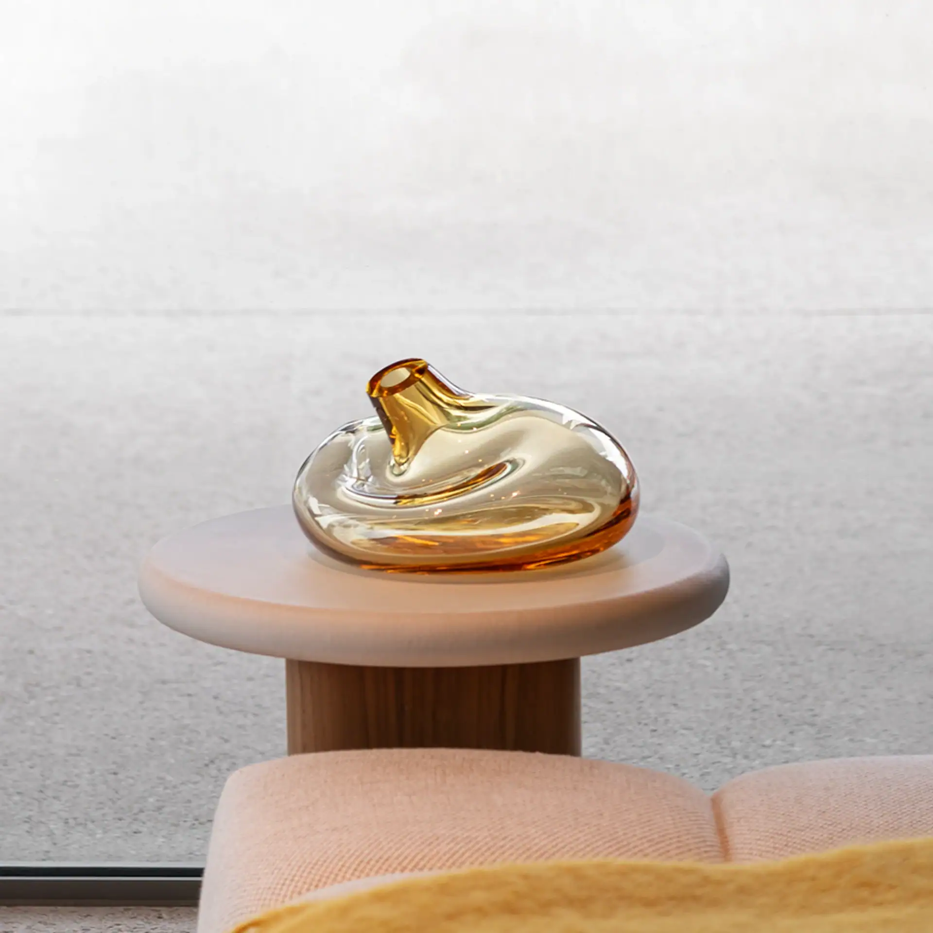 An amber glass vase with an organic, swirling shape rests on a light-colored round side table. A pink sofa and yellow blanket are in the foreground, with a light gray background.