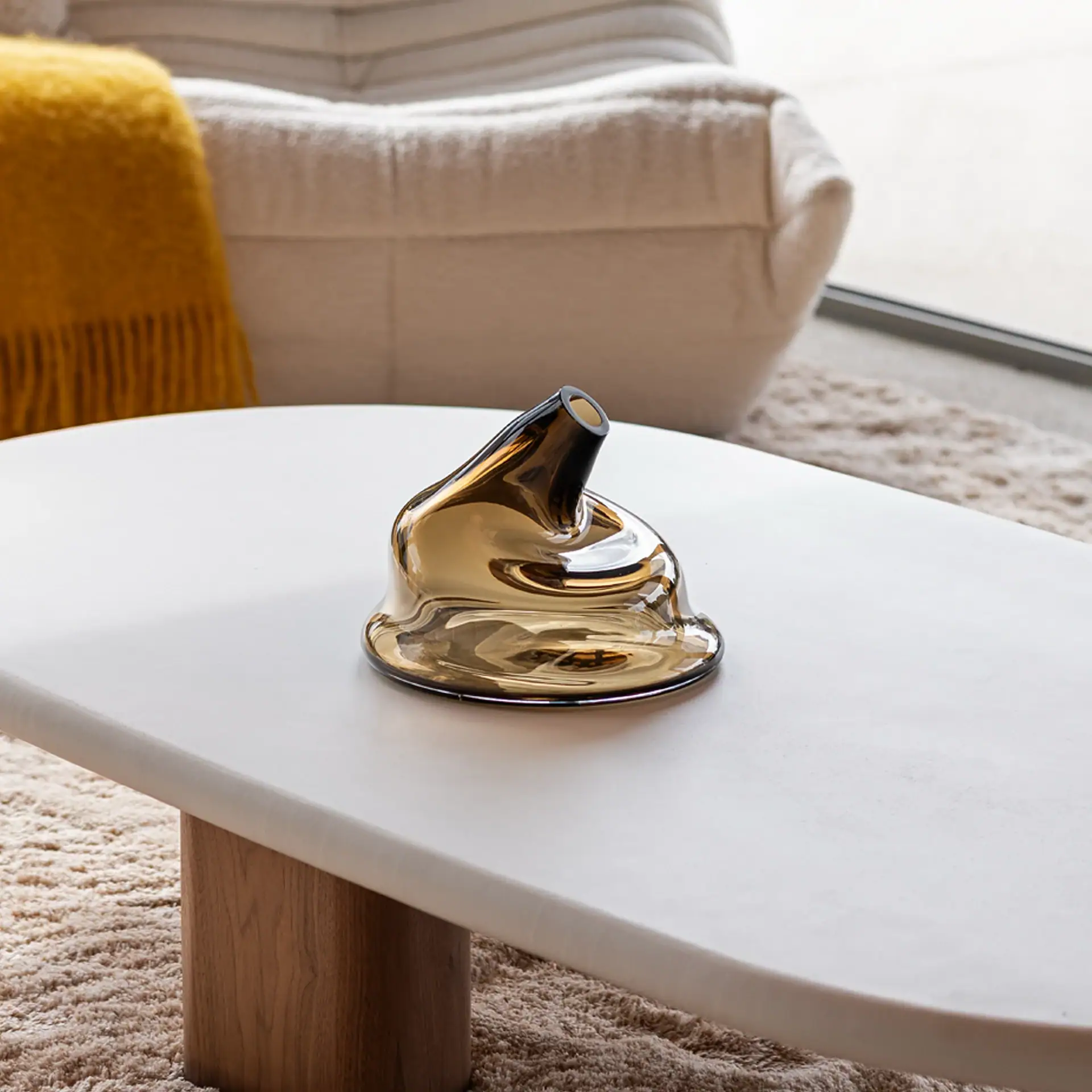 An amber-colored, irregularly shaped glass sculpture with a small opening sits on a light-colored coffee table with a wooden base, against a backdrop of a shaggy rug, a cream sofa, and a yellow throw blanket.