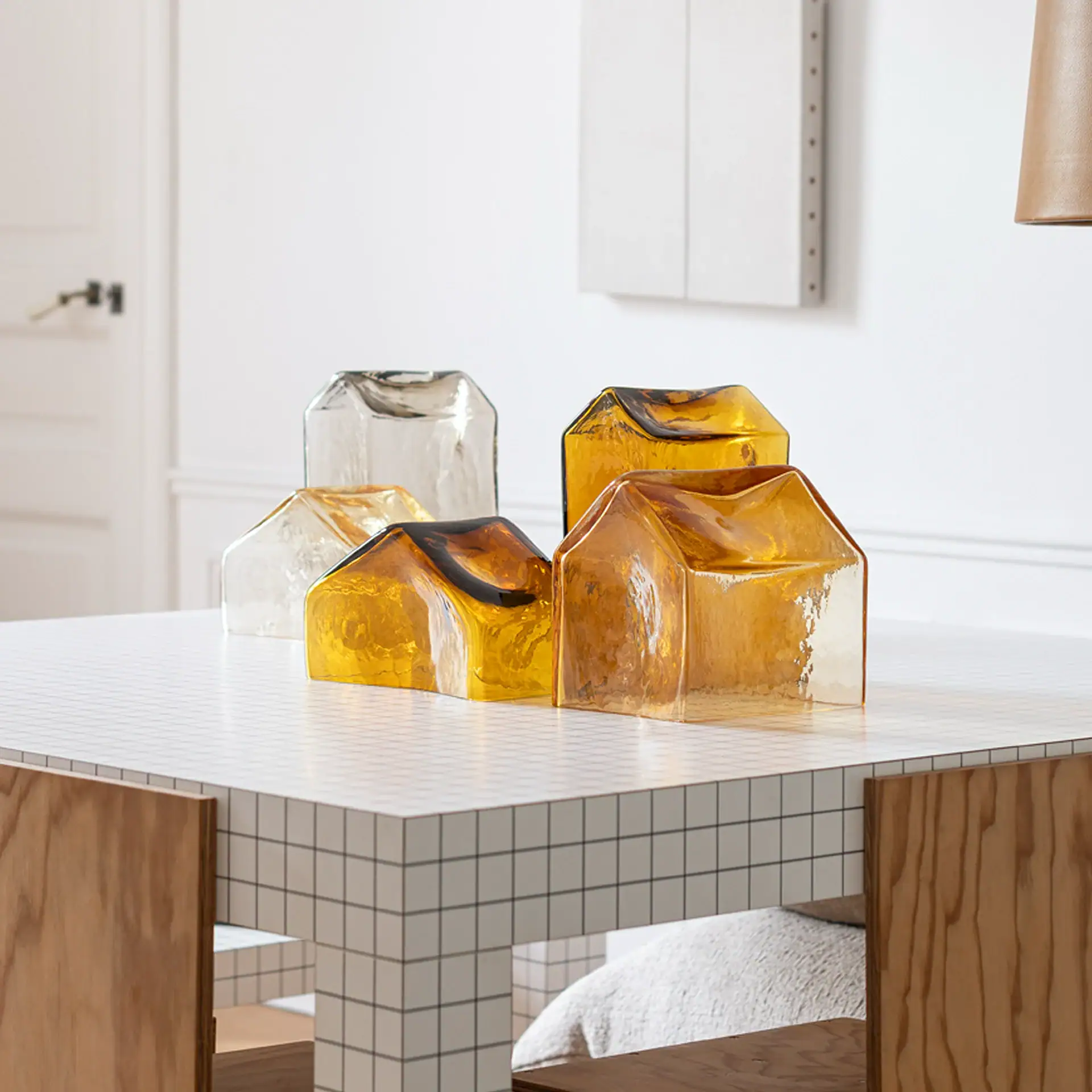Five house-shaped glass objects, two clear and three amber, on a white grid-patterned table with wooden chairs.