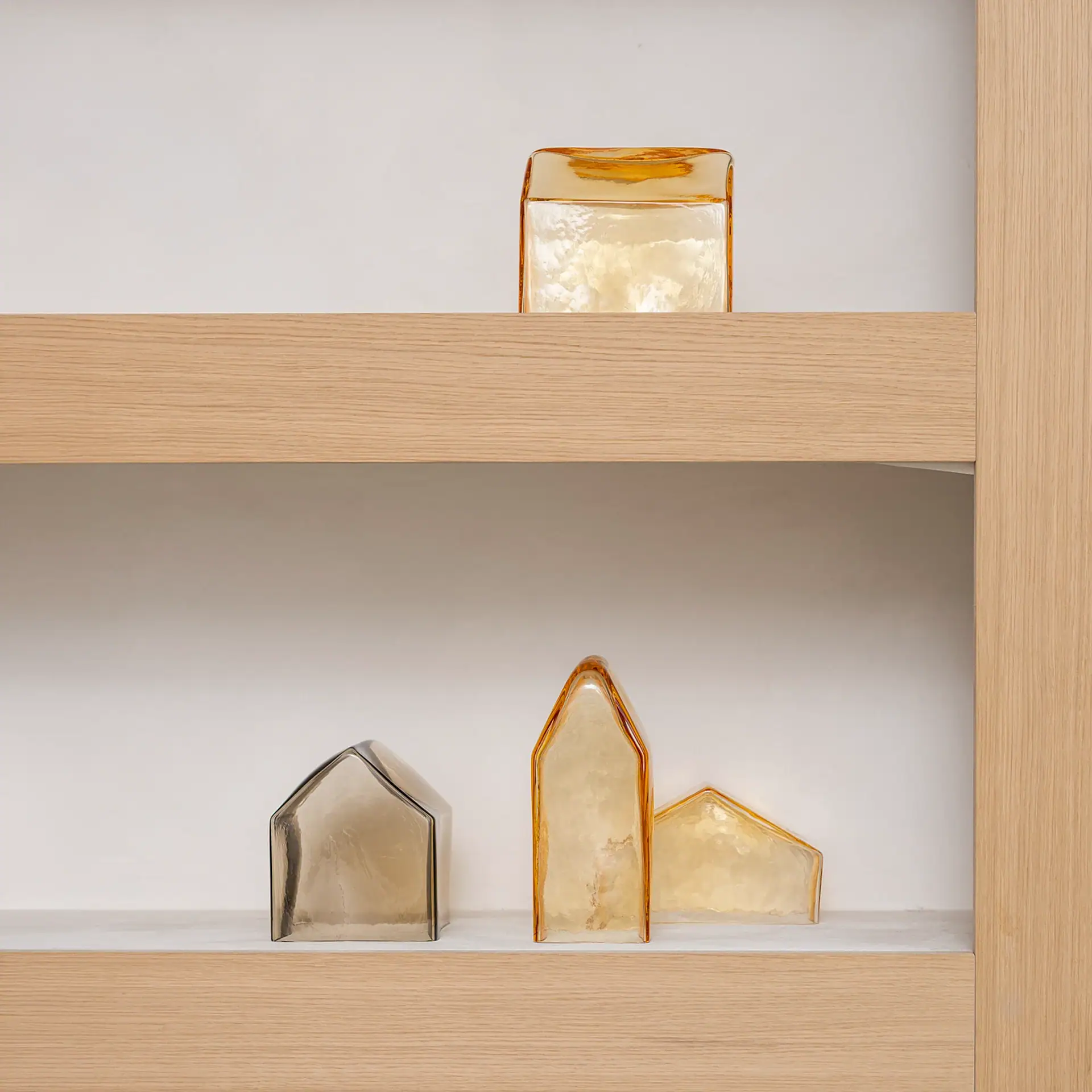 Two light wood shelves against a white wall. The top shelf holds a rectangular amber glass block. The bottom shelf displays three house-shaped glass sculptures: one grey, two amber.