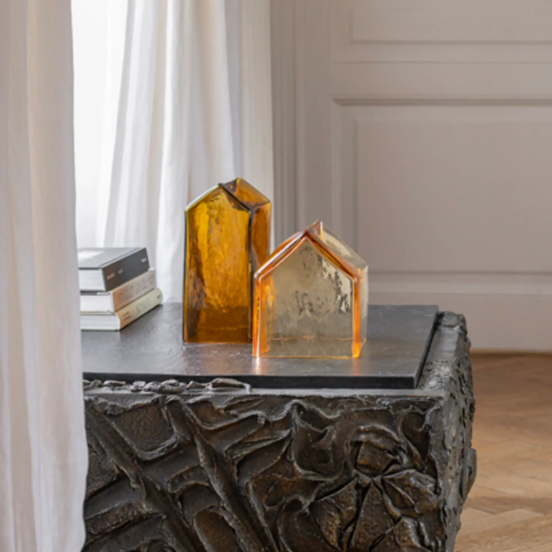 Two amber glass house-shaped objects sit on a dark, textured coffee table next to a stack of books, with white curtains and a paneled wall in the background.