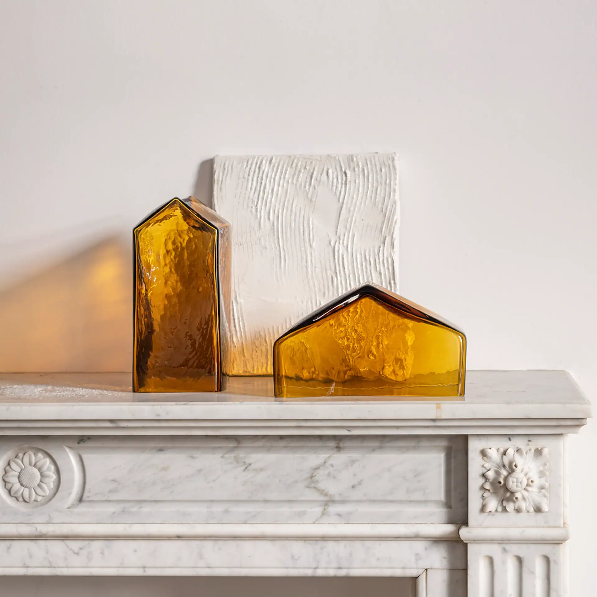 Two amber glass house-shaped objects on a white marble mantelpiece with carved details, against a white textured panel and wall.