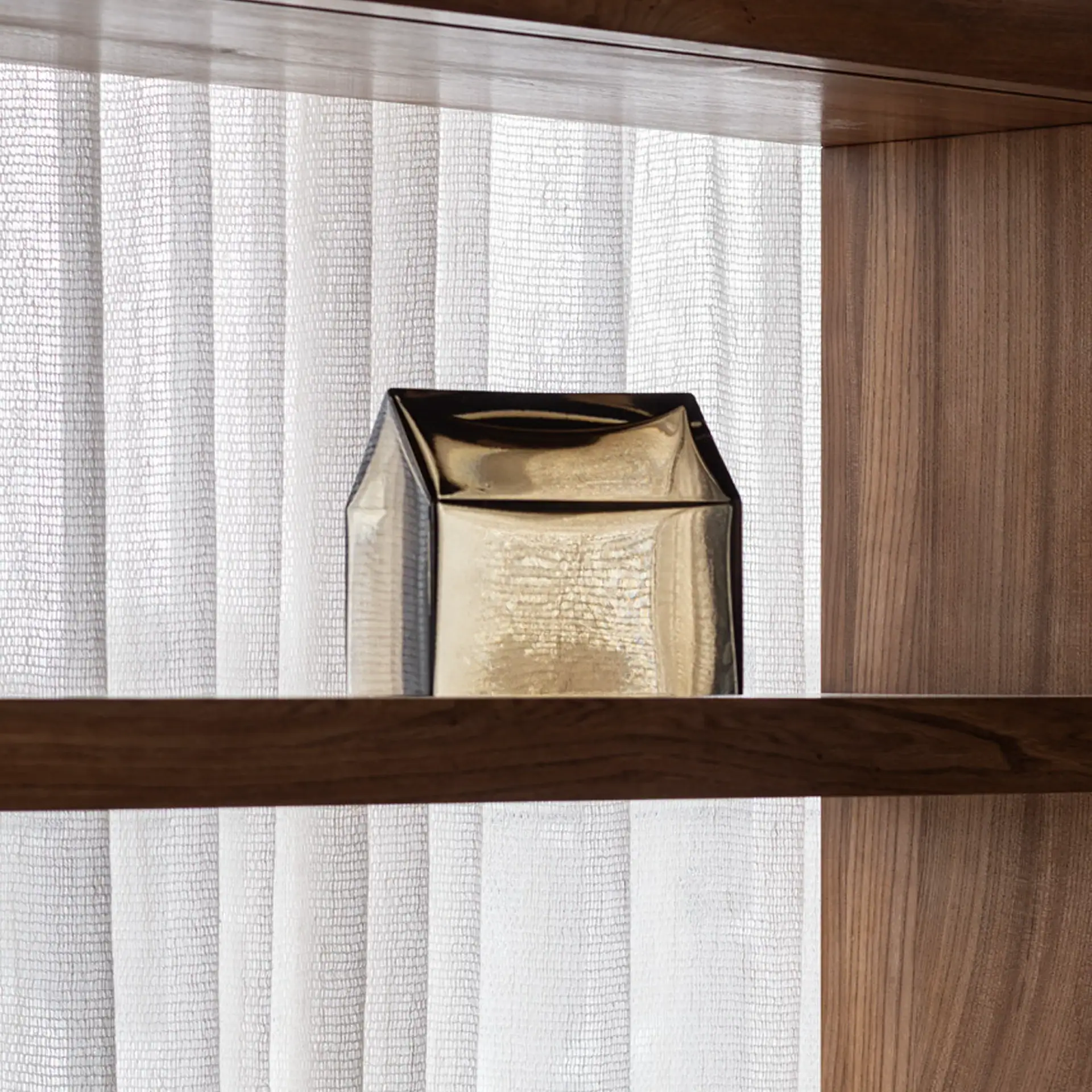A house-shaped golden-brown decorative object on a dark wooden shelf, with sheer white curtains in the background and a wooden panel to the right.