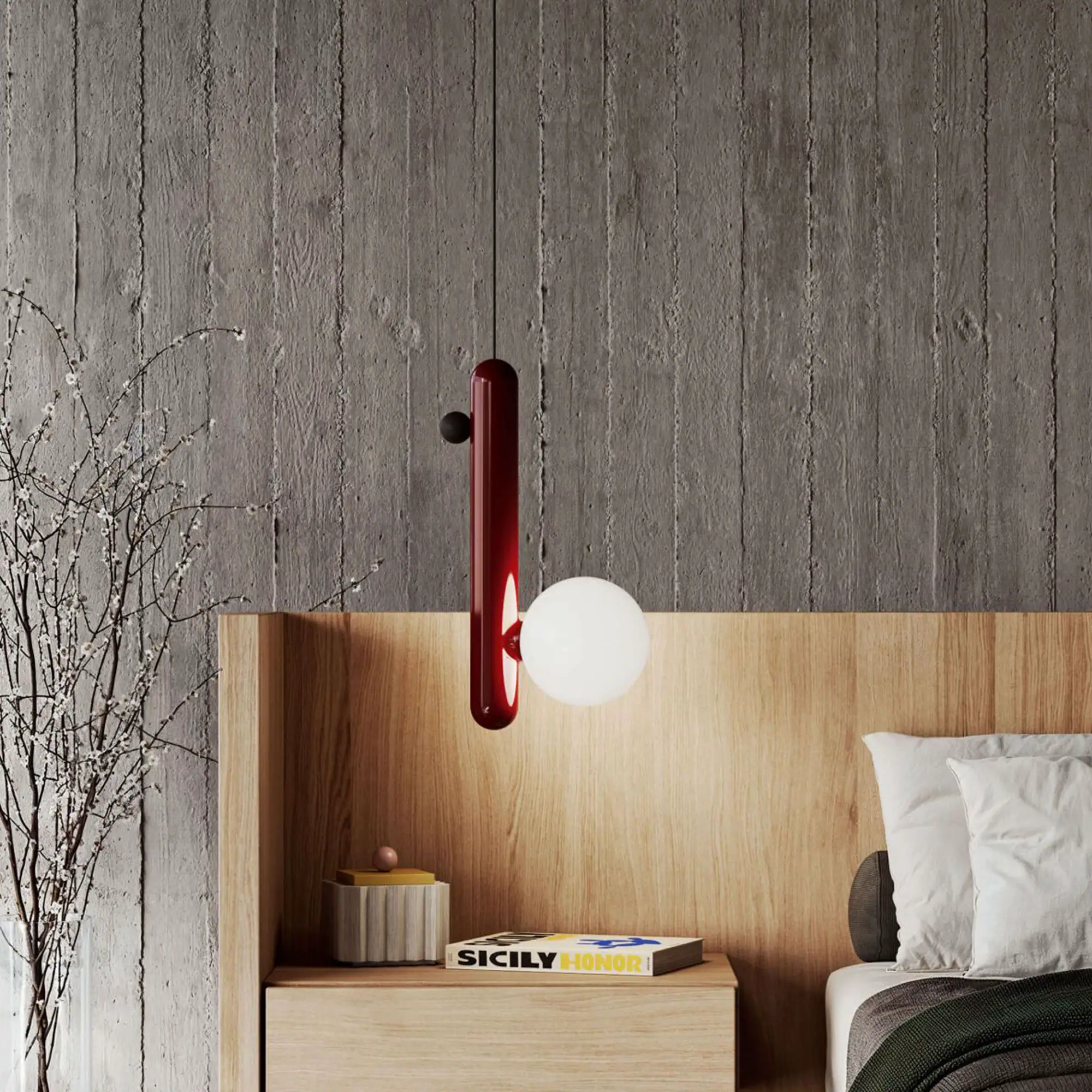 A modern bedroom with a concrete wall, wooden headboard, and a red cylindrical pendant lamp with a white spherical light. A nightstand holds a book and decor, and a vase with white blossoms is on the left. White and grey pillows are on the bed with a green blanket.