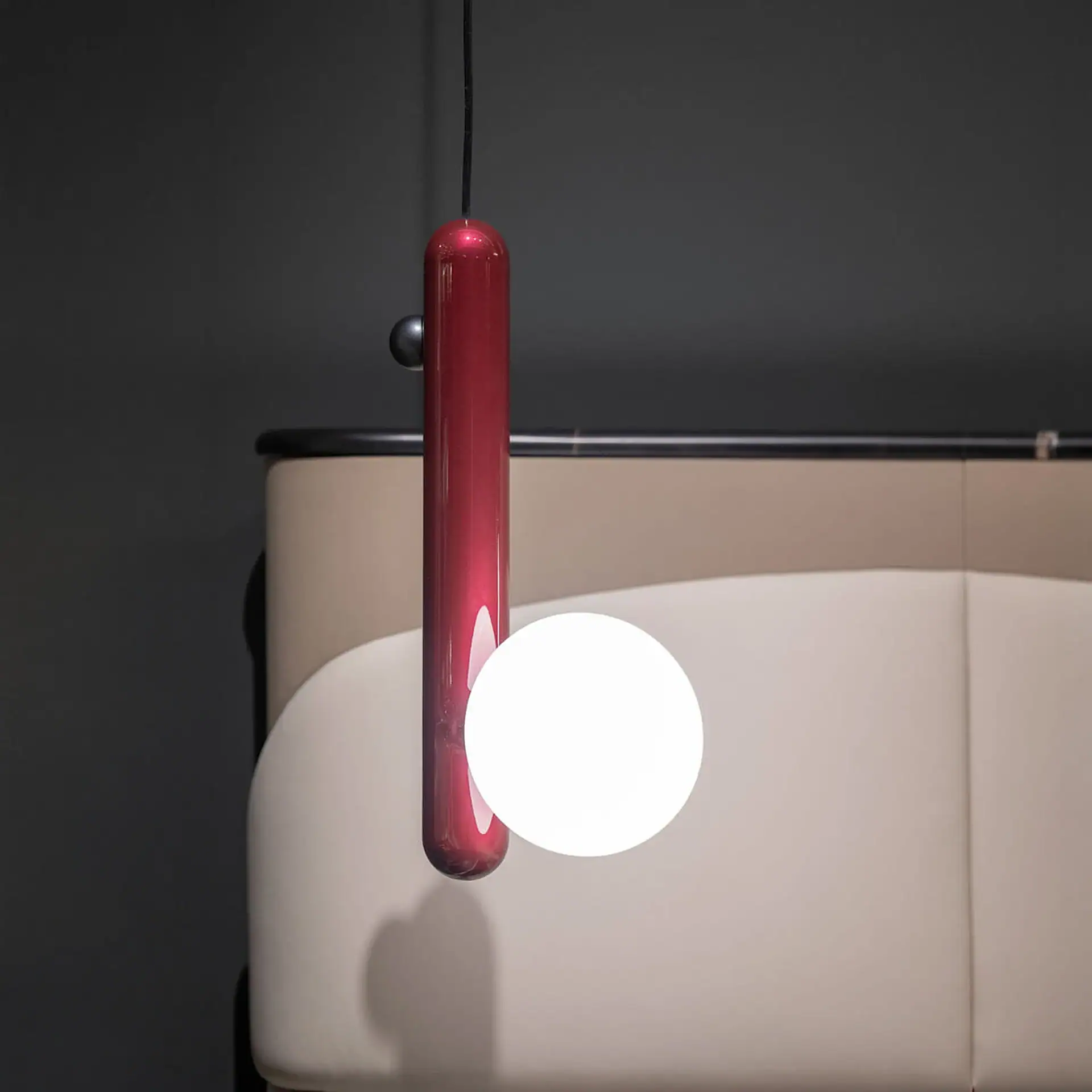 A modern pendant light with a glossy red cylindrical body and a glowing white spherical bulb, hanging against a dark wall and light beige furniture.