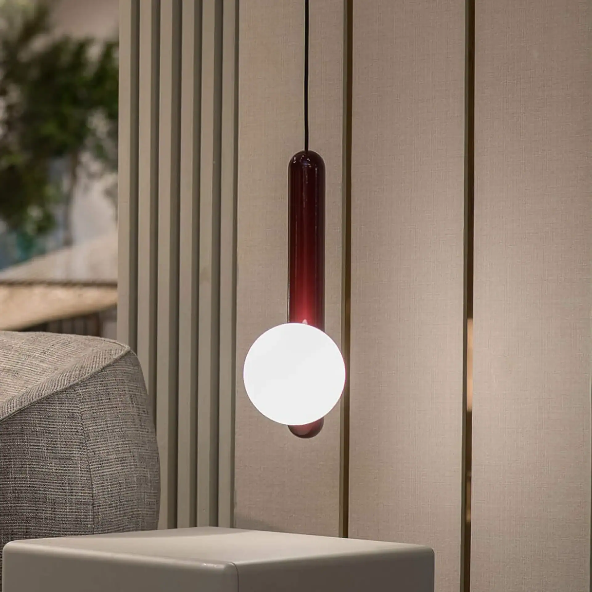 A modern pendant light with a red cylindrical top and a white spherical bulb hanging in a room with neutral-toned walls and furniture.