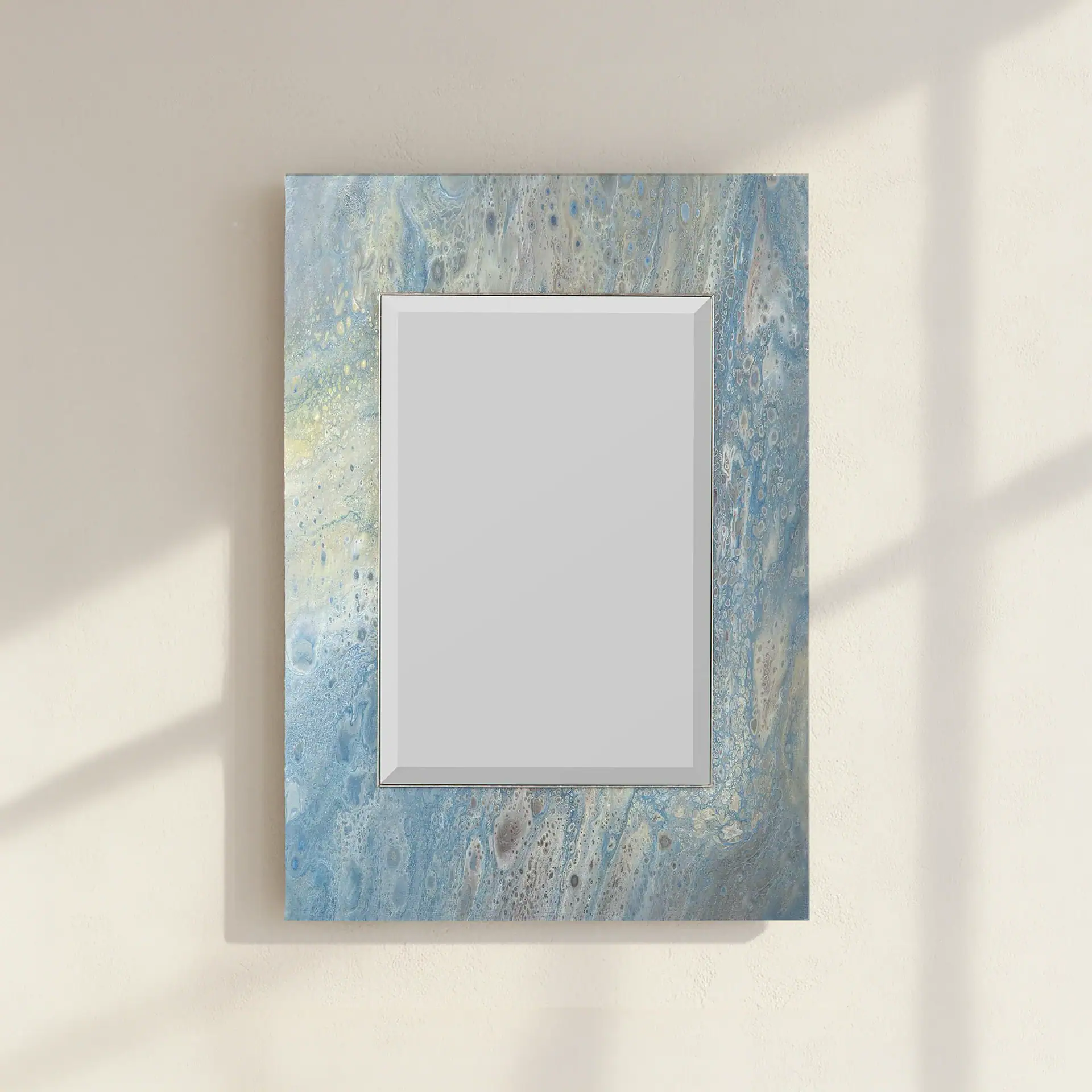 A rectangular mirror with an abstract blue, gray, and white fluid art frame hangs on a textured beige wall with soft shadows.