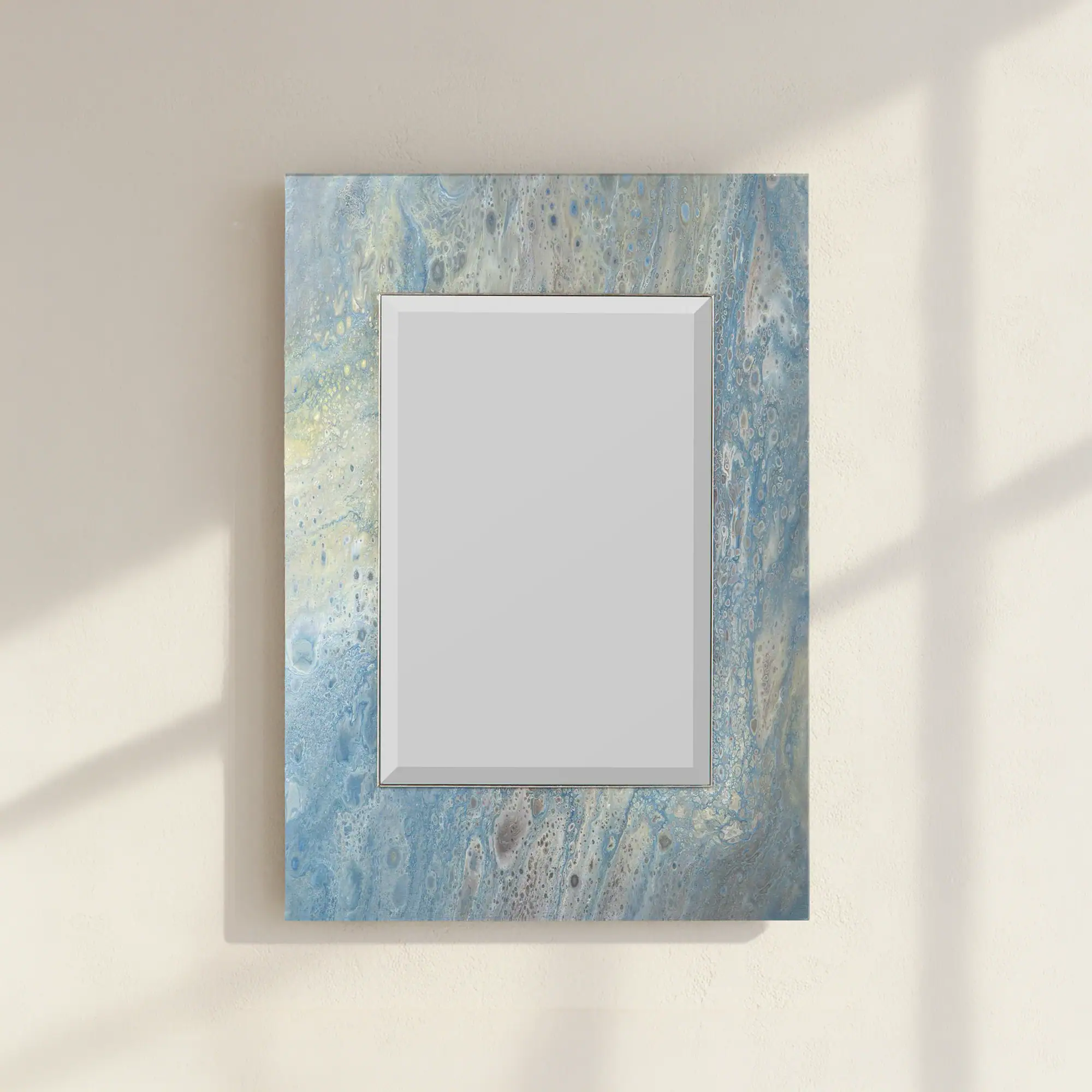 A rectangular mirror with an abstract blue, gray, and white fluid art frame hangs on a textured beige wall with soft shadows.