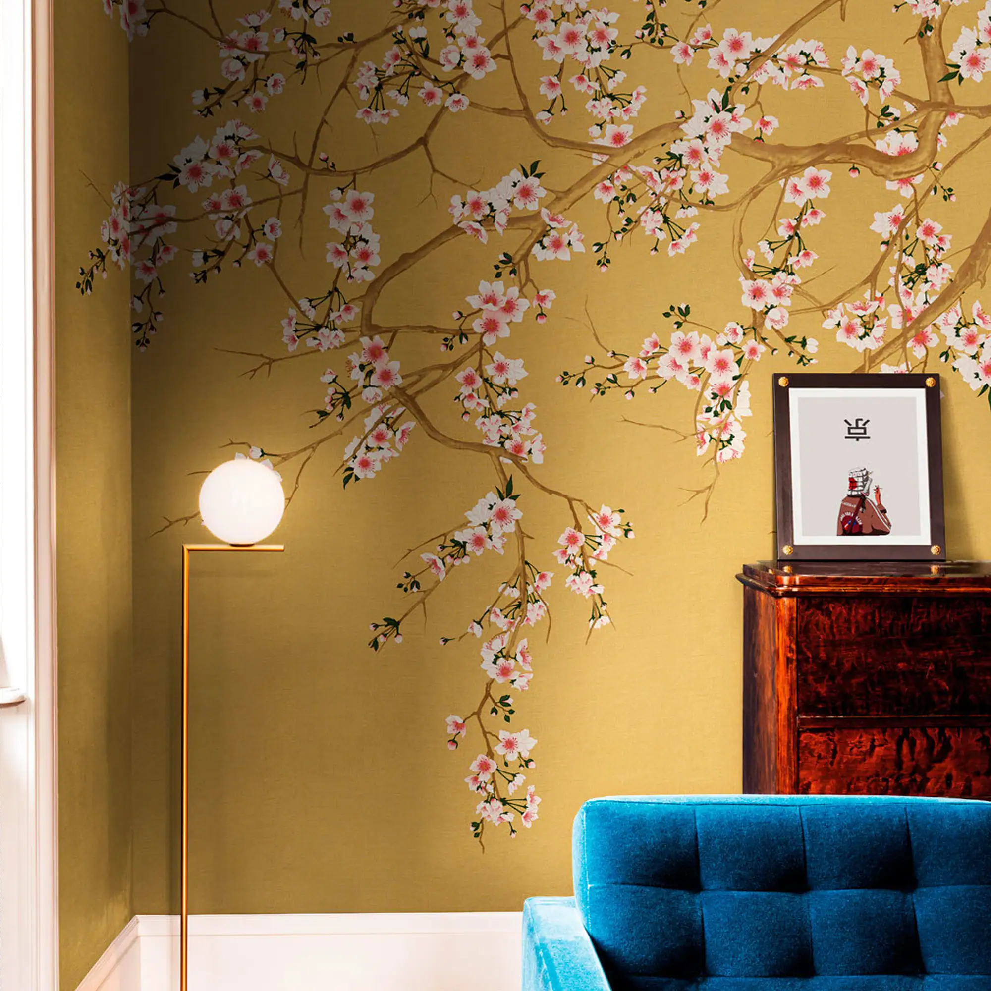 A room with a golden-yellow wall featuring a large cherry blossom mural. A modern floor lamp, a dark wooden cabinet with a framed picture, and a blue tufted sofa are visible.